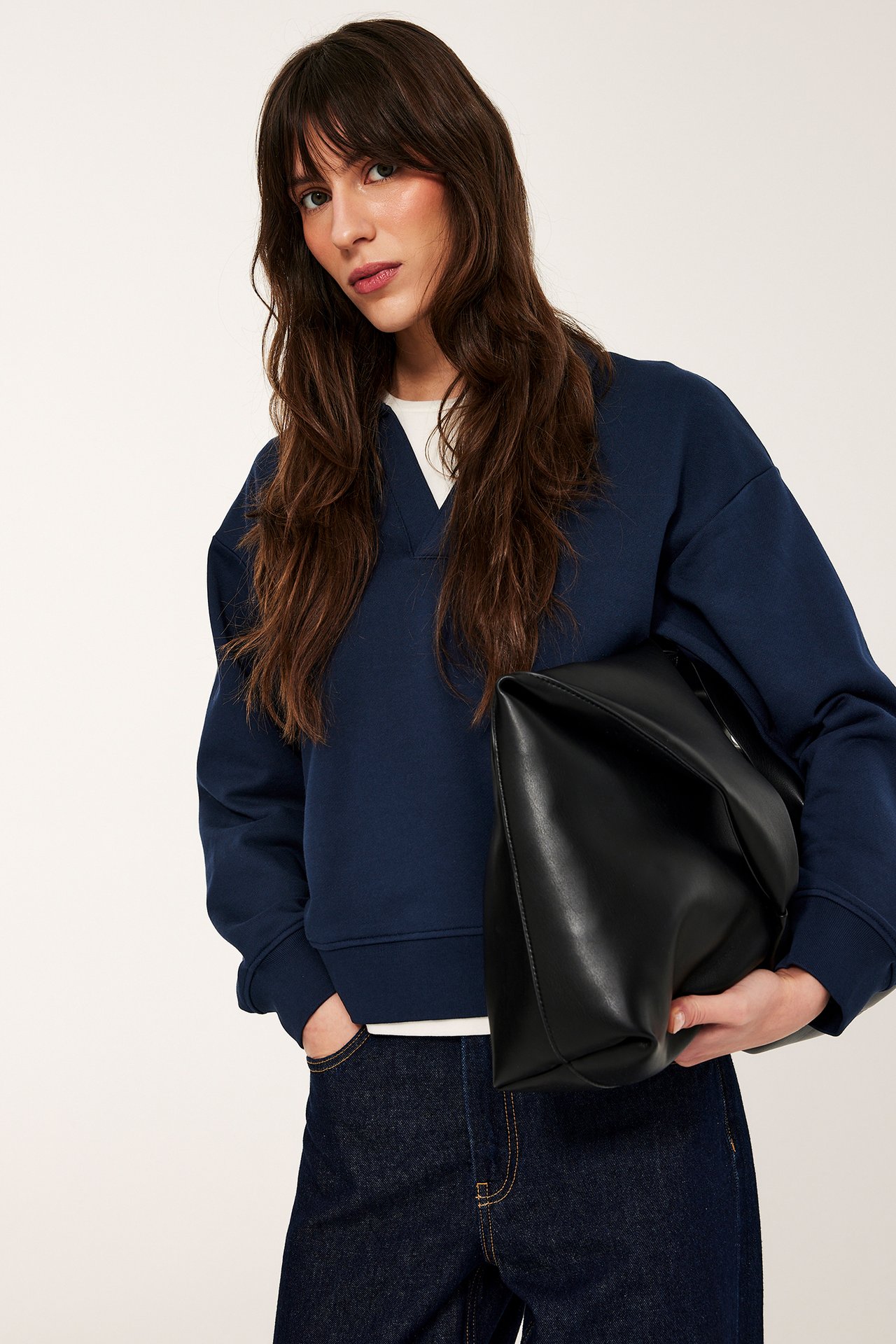 Nainen tummansinisessä V-kaula-aukkopuserossa valkoisen topin päällä, tummissa farkuissa, pitää mustaa käsilaukkua.