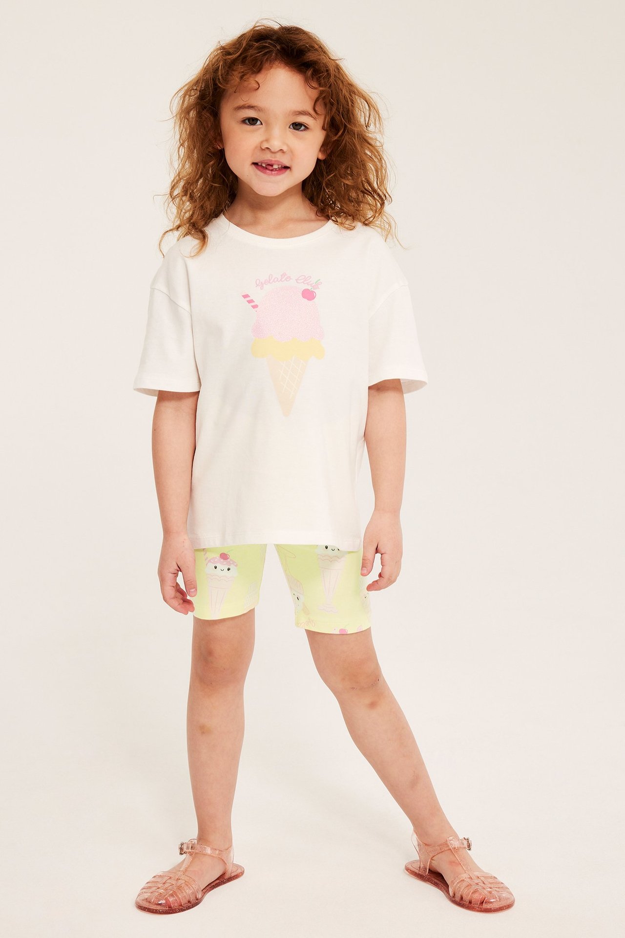 Edestä: Lapsi käyttää vaaleankeltaisia jäätelökuvioisia pyöräilyshortseja, valkoista t-paitaa ja vaaleanpunaisia sandaaleita.