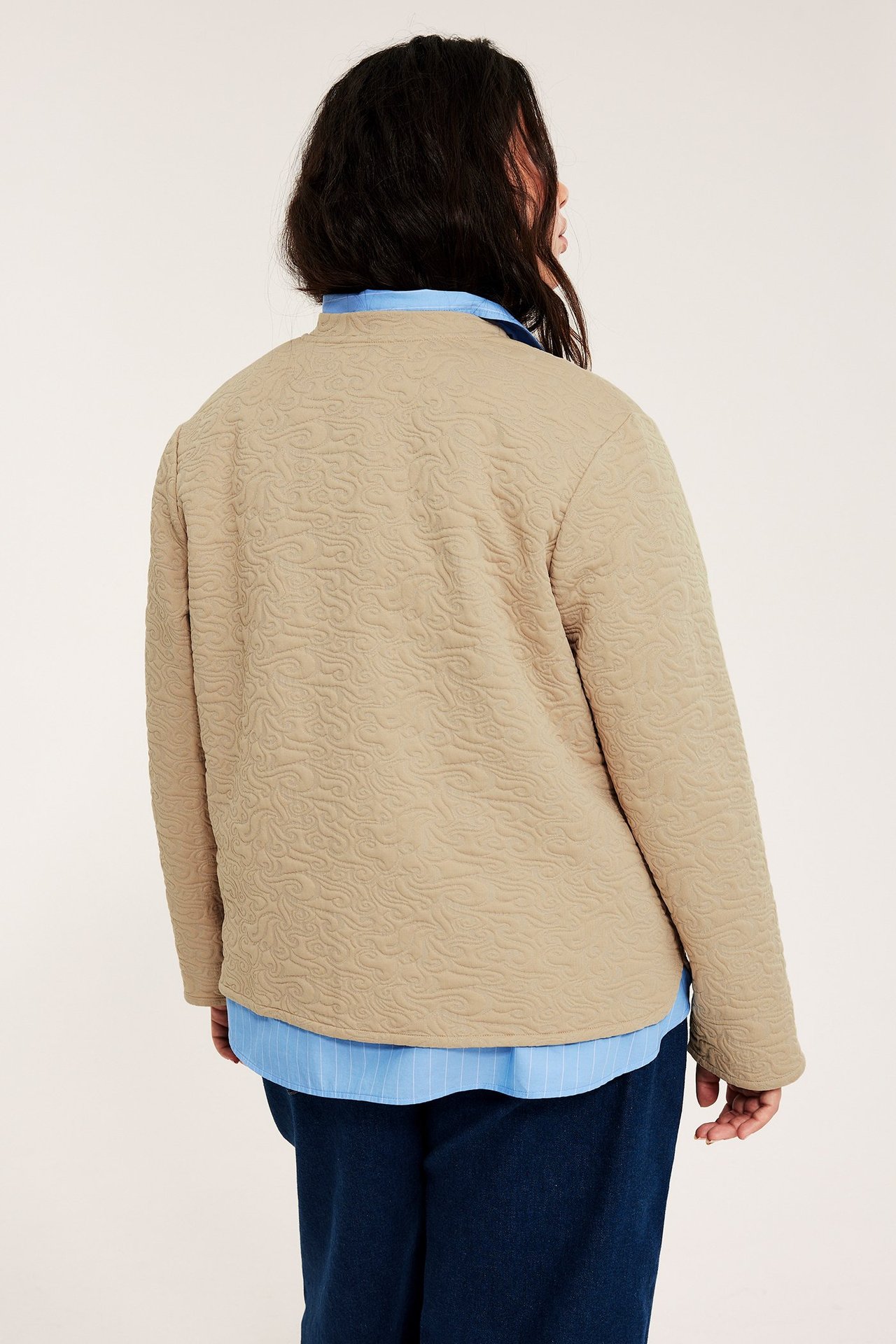 Naisen takakuva: beige tikattu takki vaaleansinisen paidan ja tummansinisten housujen päällä.