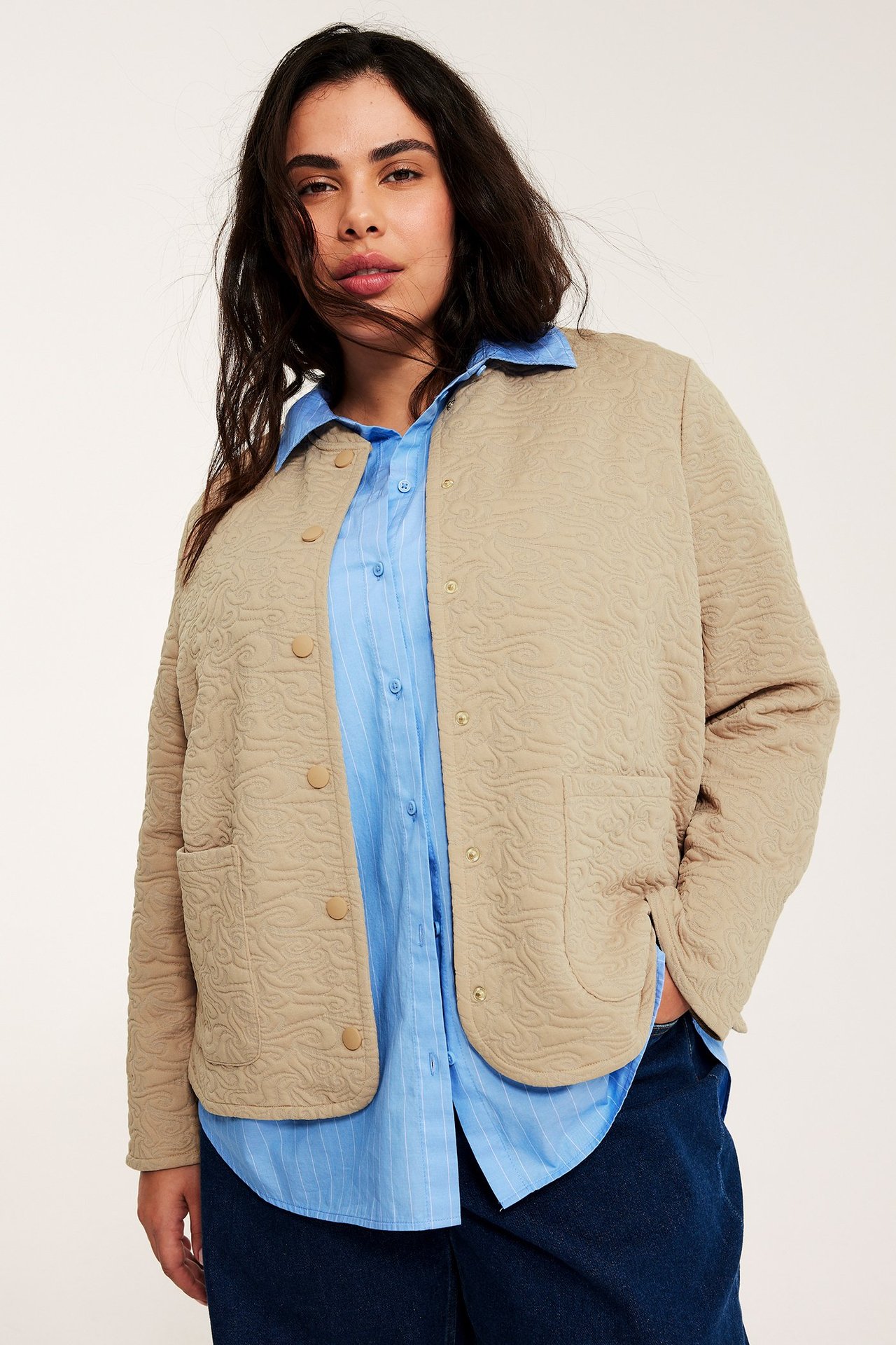 Frontvy: Kvinna i öppen beige quiltad jacka med vågmönster, buren över en blårandig skjorta.