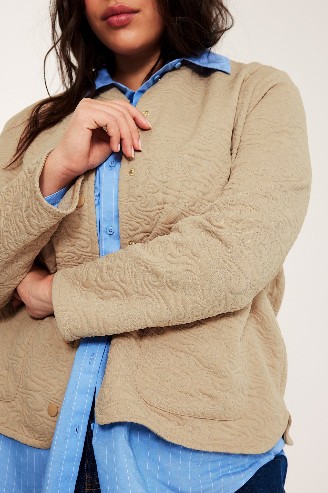 Lähikuva edestä: nainen beigessä tikatussa takissa, neppareilla, vaaleansinisen raidallisen paidan päällä.