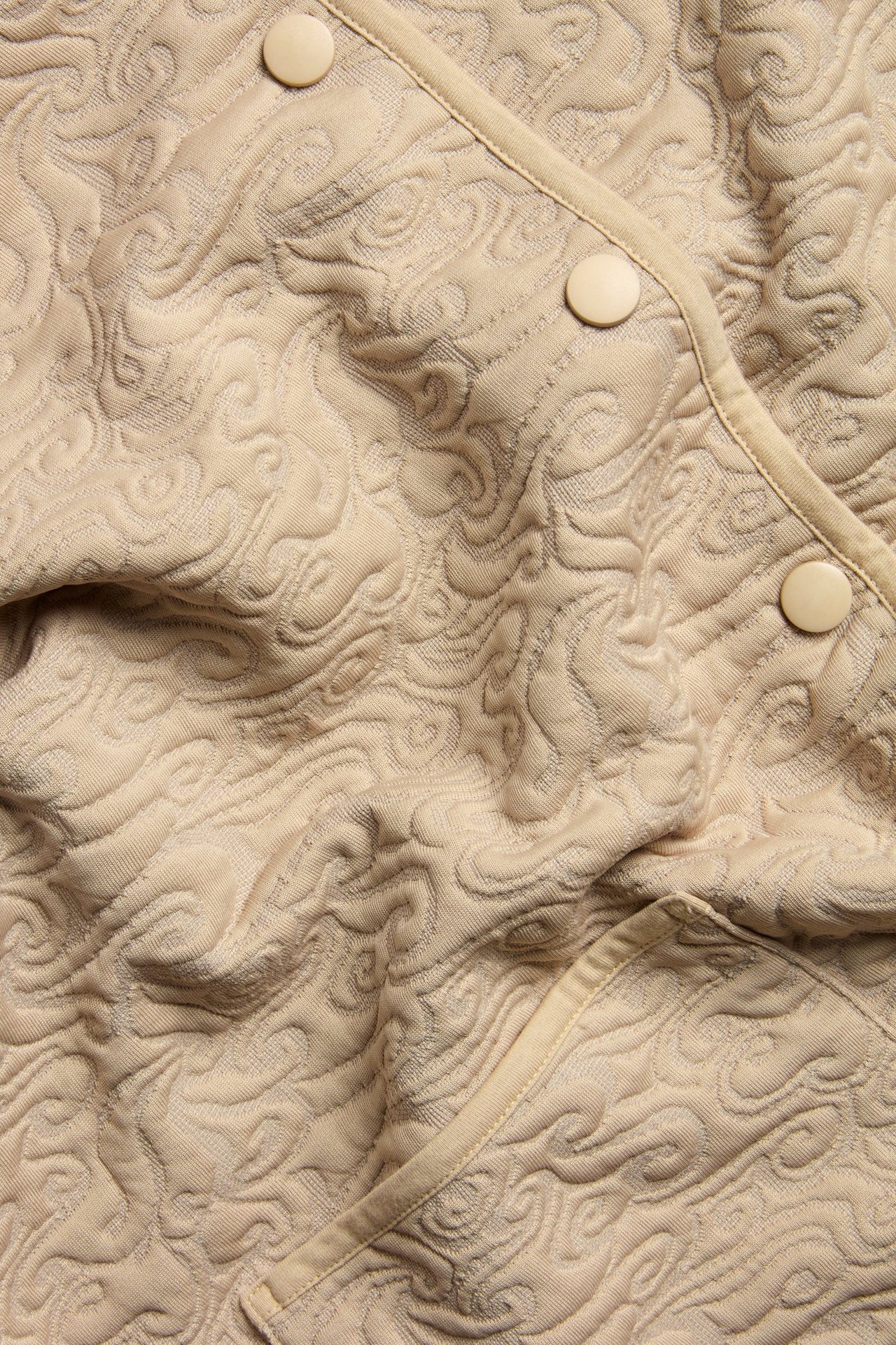 Nærbilde av en beige quiltet jakke med preget mønster og tre synlige runde knapper.