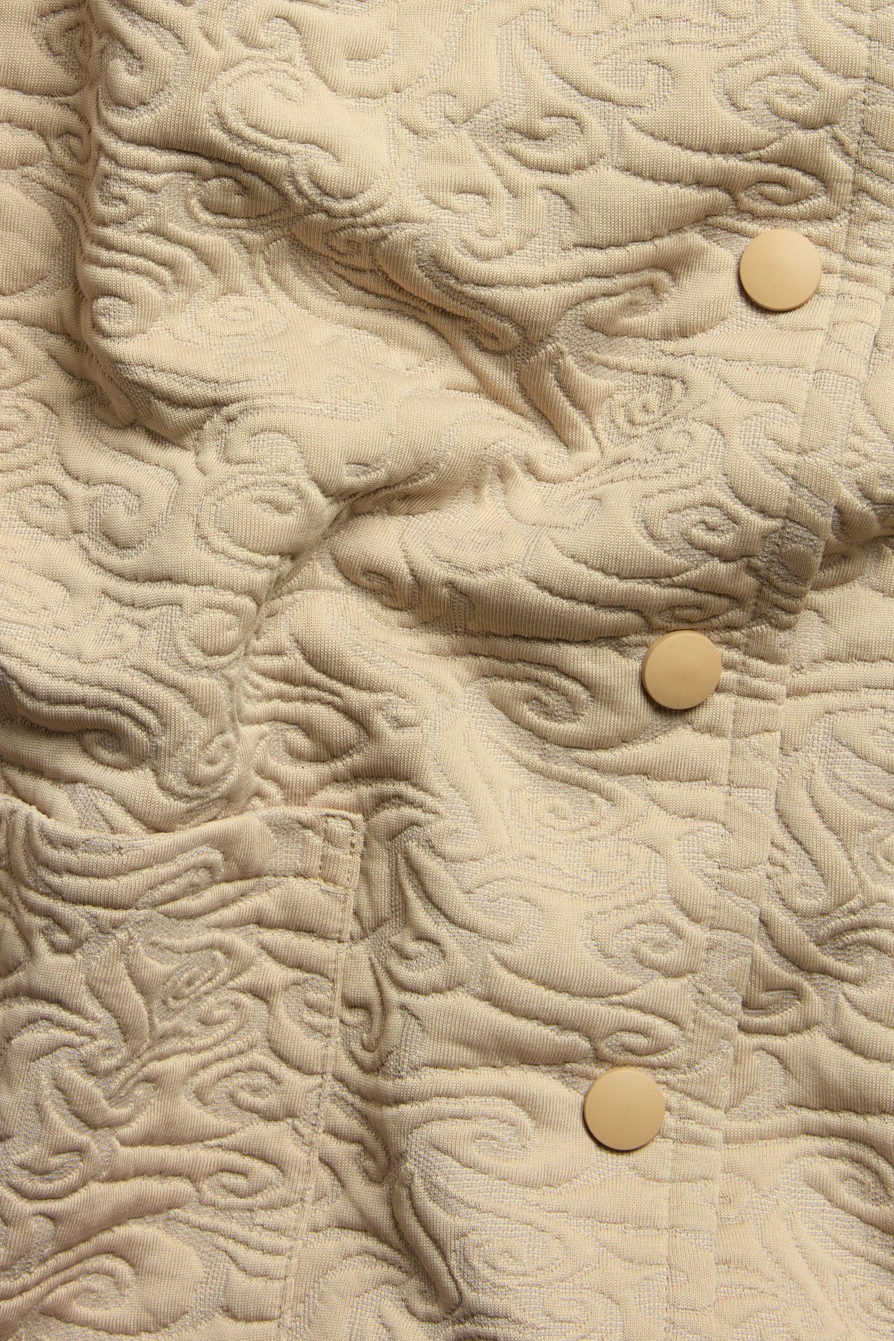 Zbliżenie beżowej pikowanej kurtki z wirującym wzorem i trzema okrągłymi guzikami.