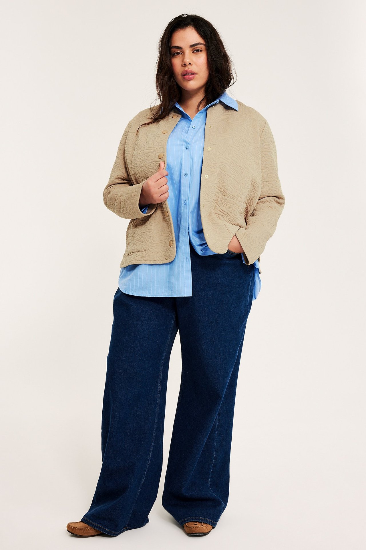 Kvinna bär en beige quiltad jacka, blårandig skjorta, mörkblå vida jeans och bruna skor.