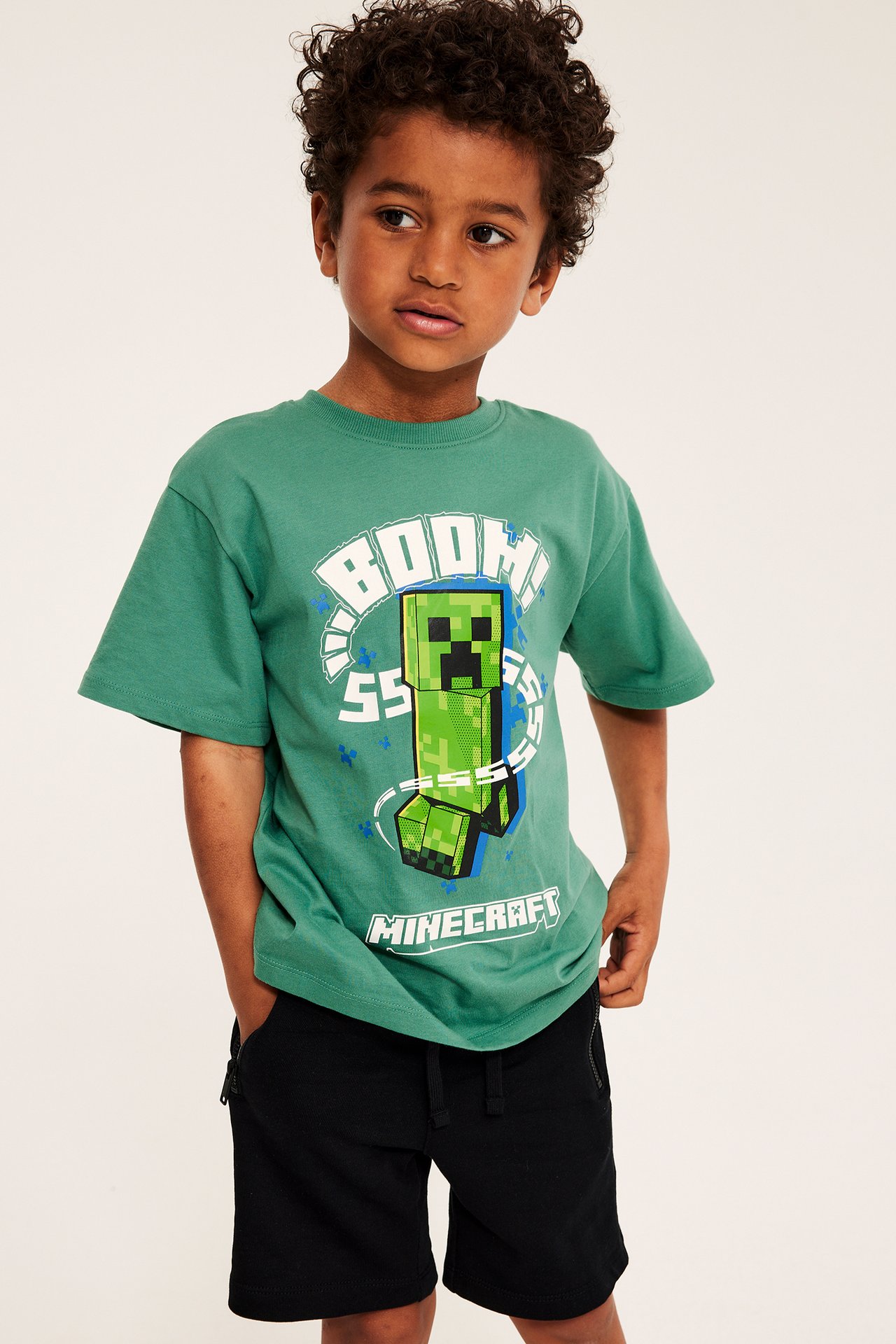 Framifrån: Ett barn i grön oversized bomulls-T-shirt med Minecraft-tryck och svarta dragsko-shorts.