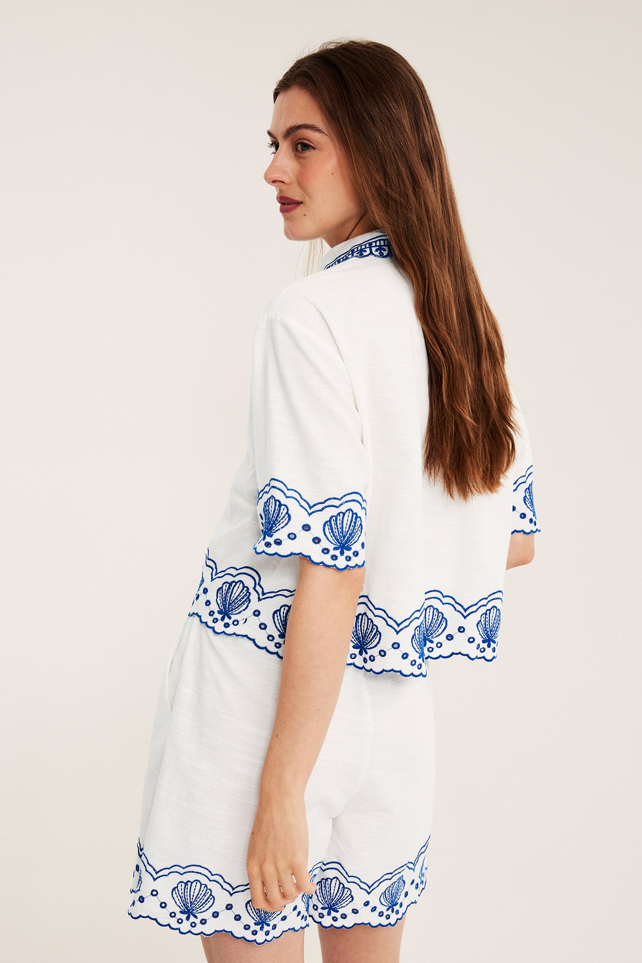 Sidebilde av kvinne i hvit kortermet bomullsskjorte og matchende shorts med blå skjellbroderi.