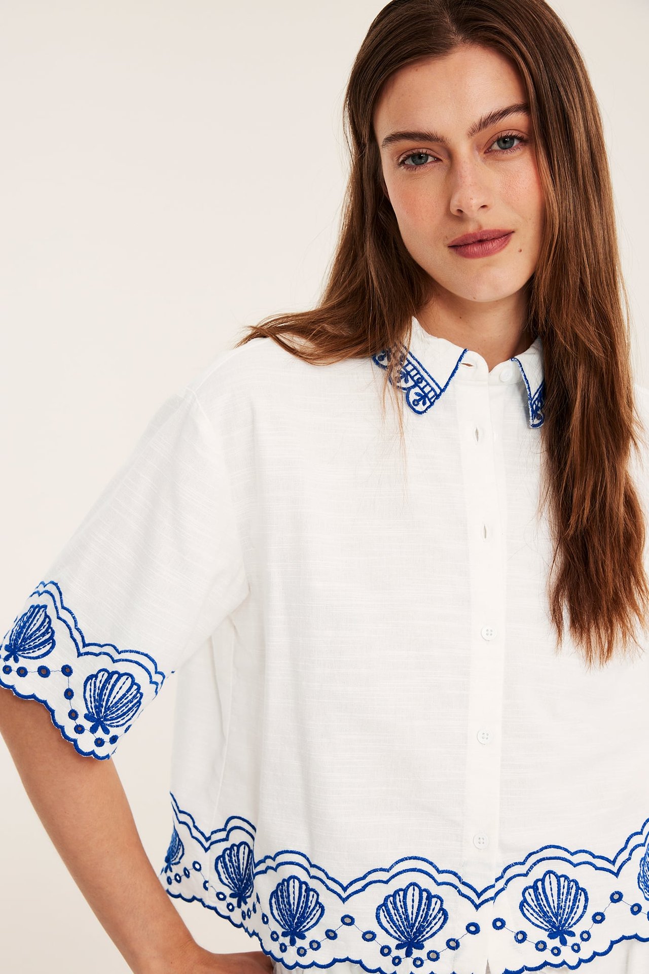 Närmare frontbild: Kvinna bär vit kortärmad bomullsskjorta med blått snäckskalsbroderi på krage, ärmar och nederkant.