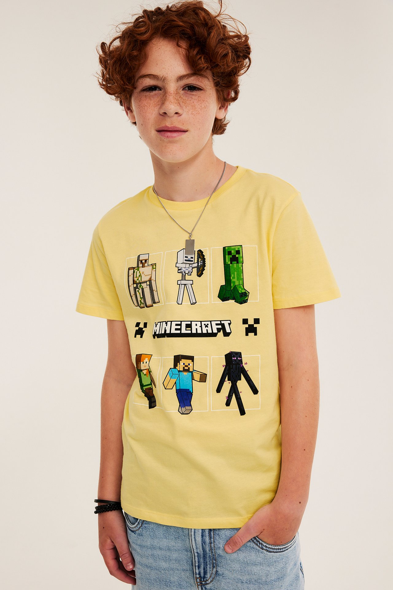 Frontbilde av et barn i gul bomulls-T-skjorte med Minecraft-trykk, lyseblå jeans, sølvkjede og svart armbånd.