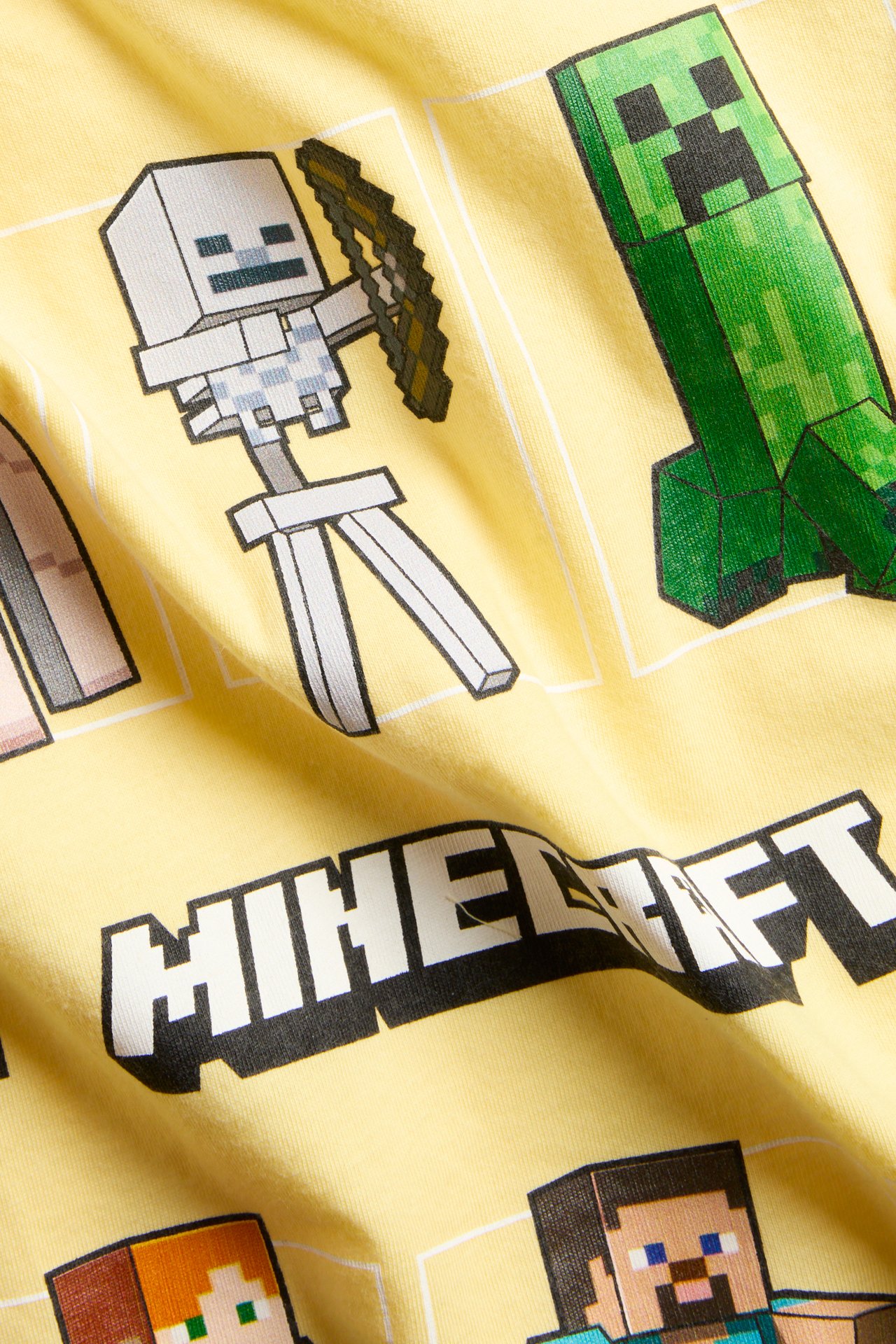 Zbliżenie na żółtą bawełnianą koszulkę dziecięcą z kolorowym nadrukiem postaci Minecraft i logo z przodu.