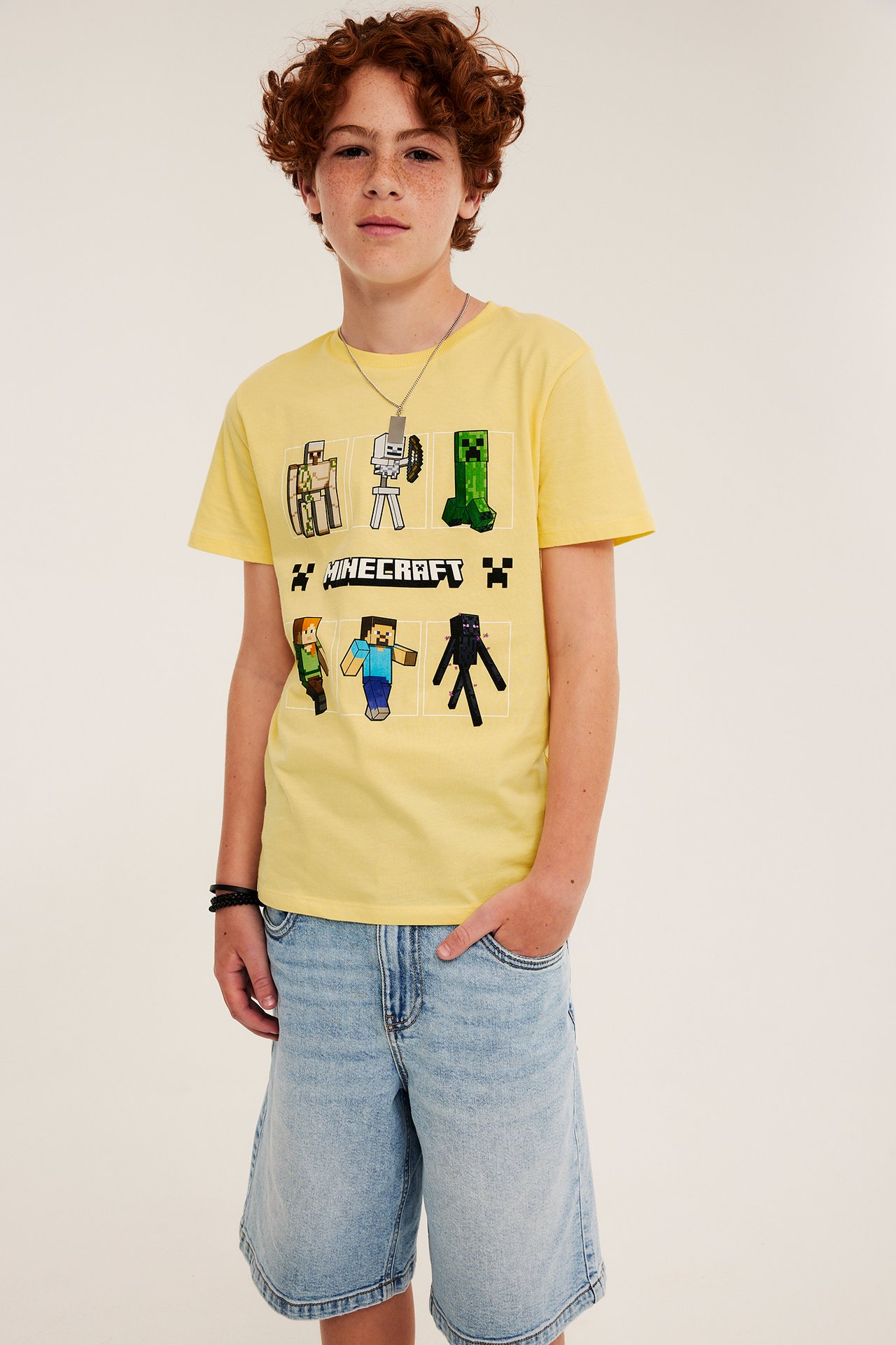 Forfra: barn i gul bomulls-Minecraft-T-skjorte, lyseblå dongerishorts, sølvkjede og svart armbånd.