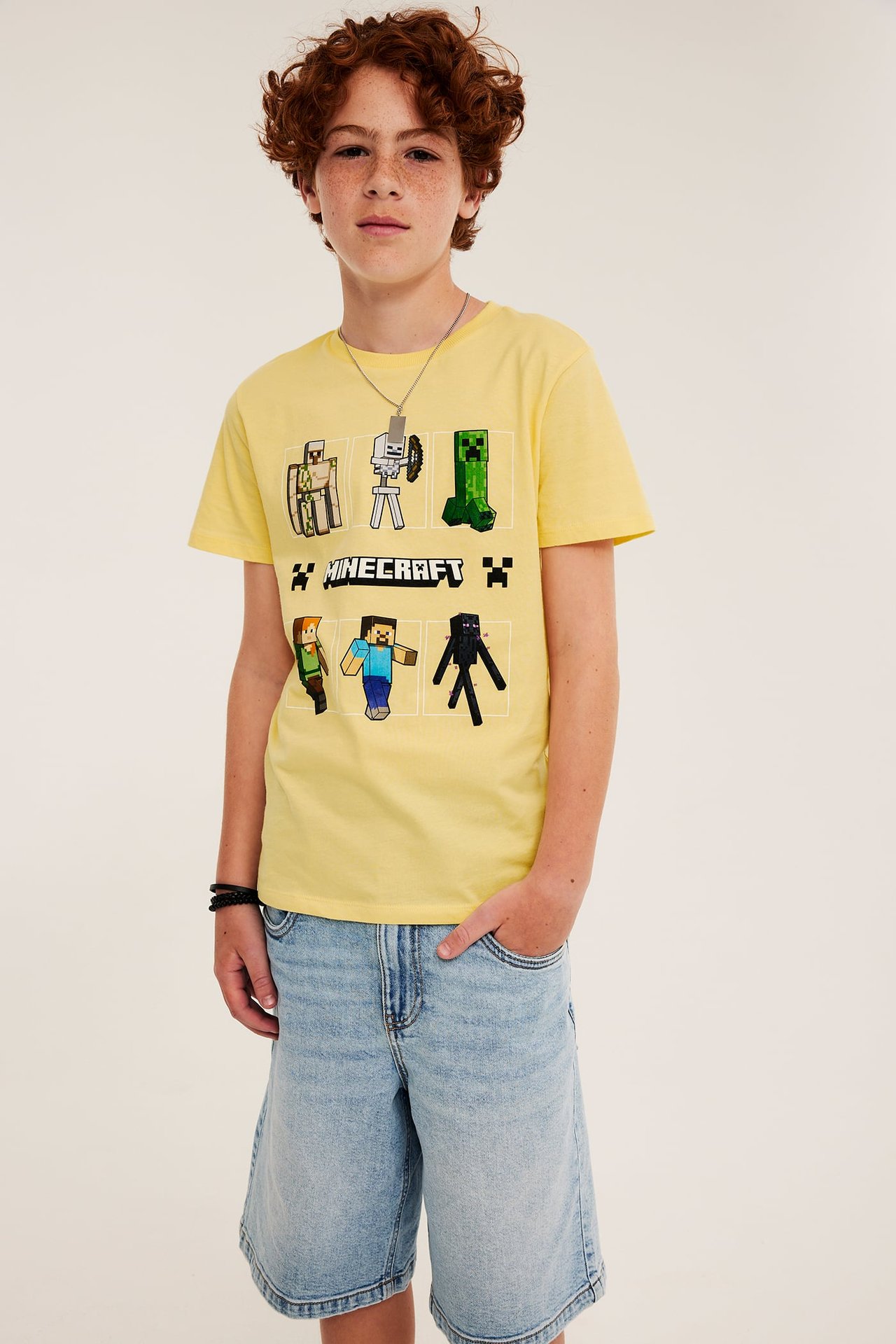 Forfra: barn i gul bomulls-Minecraft-T-skjorte, lyseblå dongerishorts, sølvkjede og svart armbånd.