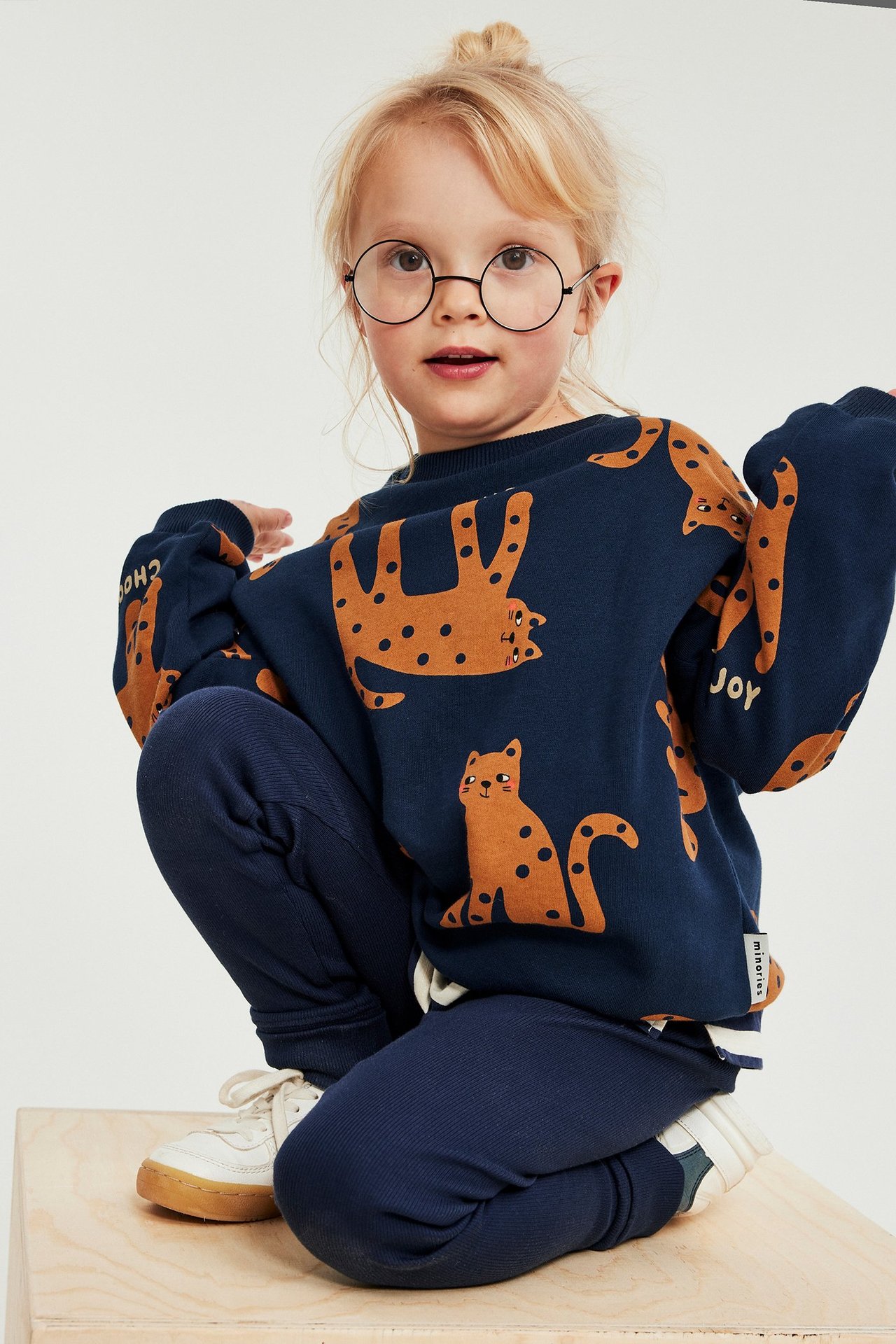 Lapsi edestä: tummansininen puuvillapusero oranssilla kissaprintillä, tummansiniset leggingsit ja valkoiset tennarit.