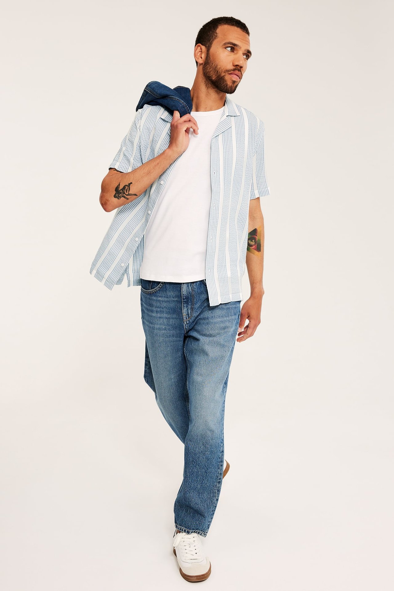 Framifrån: Man i ljusblå randig kortärmad bäckeböljaskjorta, vit t-shirt, blå jeans och vita sneakers.