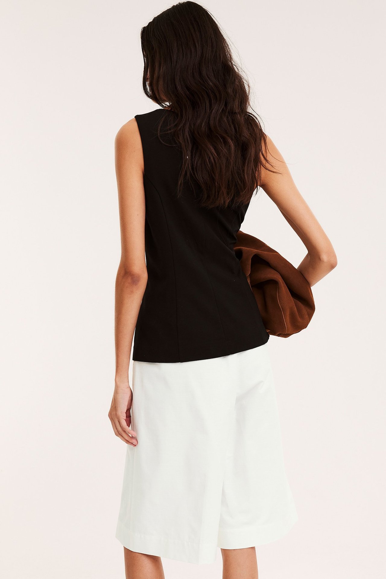 Naisen selkäpuoli valkoisissa puuvillasekoite-bermudashortseissa, mustassa hihattomasa topissa ja ruskea vaate käsivarrella.