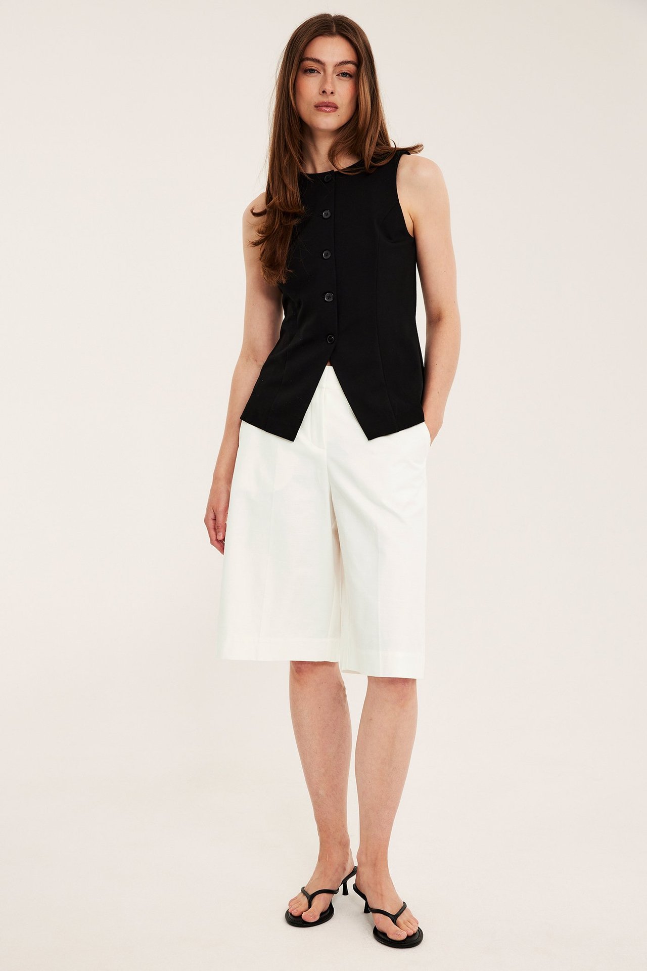 Frontvisning: kvinne i hvite bomullsmix bermudashorts, svart ermeløs topp, svarte heelflip-flops.