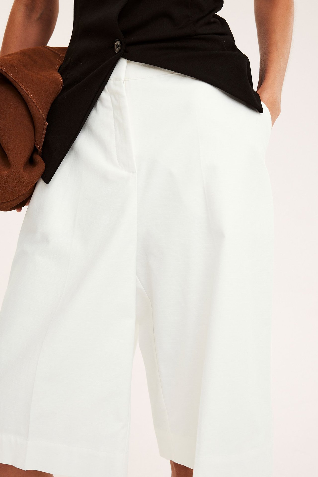 Nærbilde av en kvinne i hvite knelange bermudashorts, svart ermeløs topp og en drapert brun jakke.