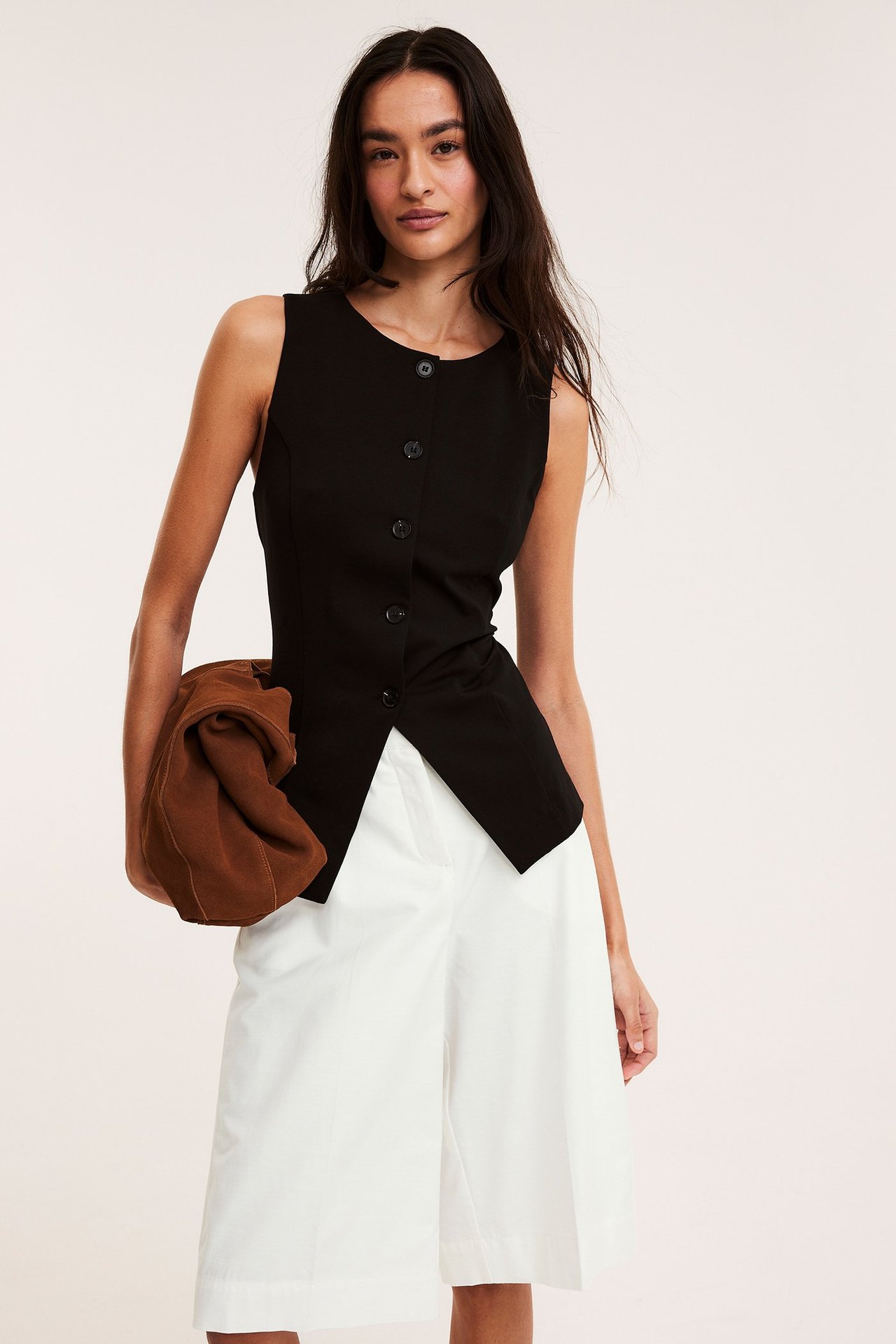 Forfra: kvinne i hvite bomullsbermudashorts, svart knappet ermeløs topp, holder en brun veske.