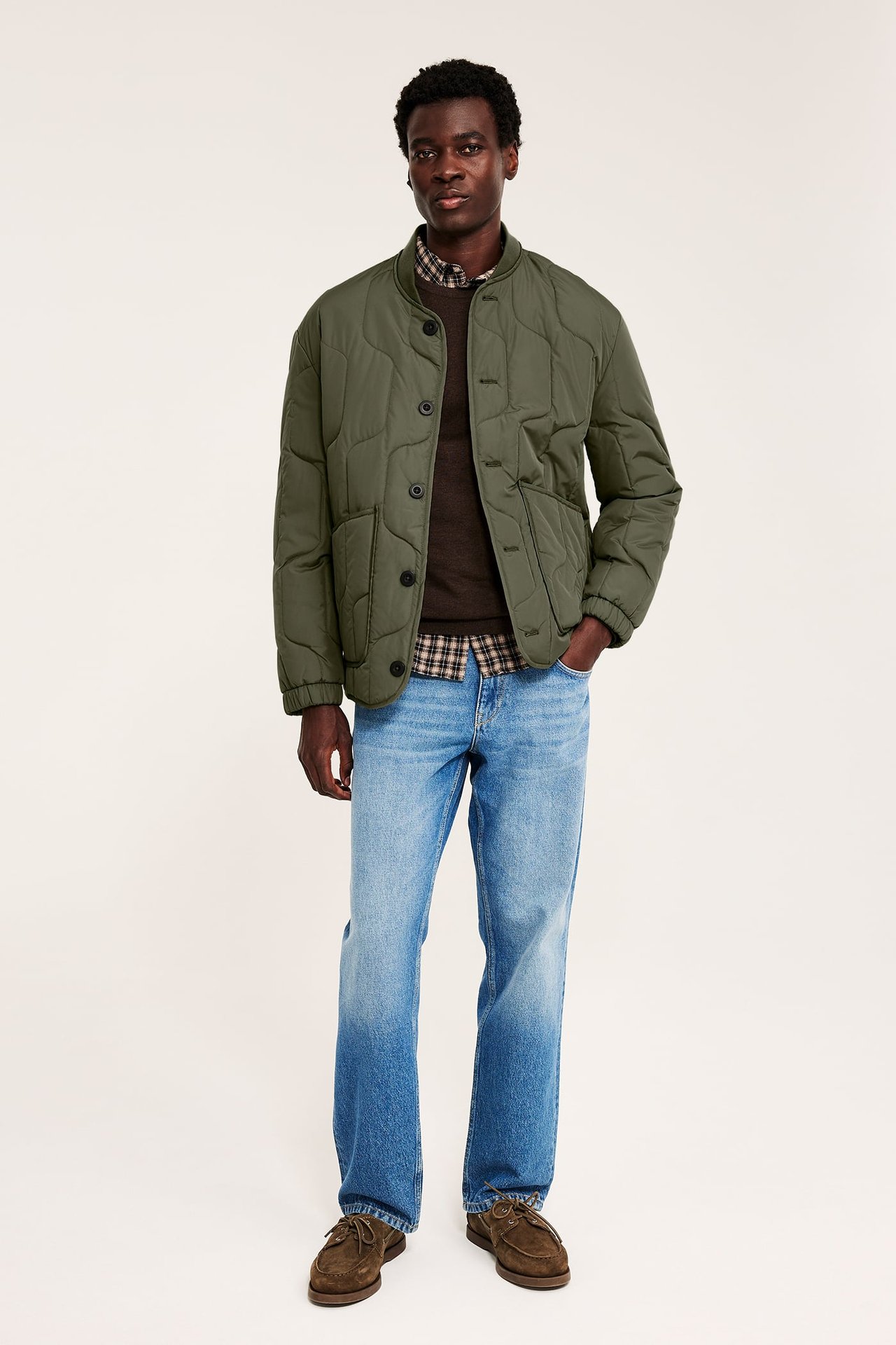 Frontbilde av mann i grønn vattert linerjakke over brun genser, blå jeans og brune sko.