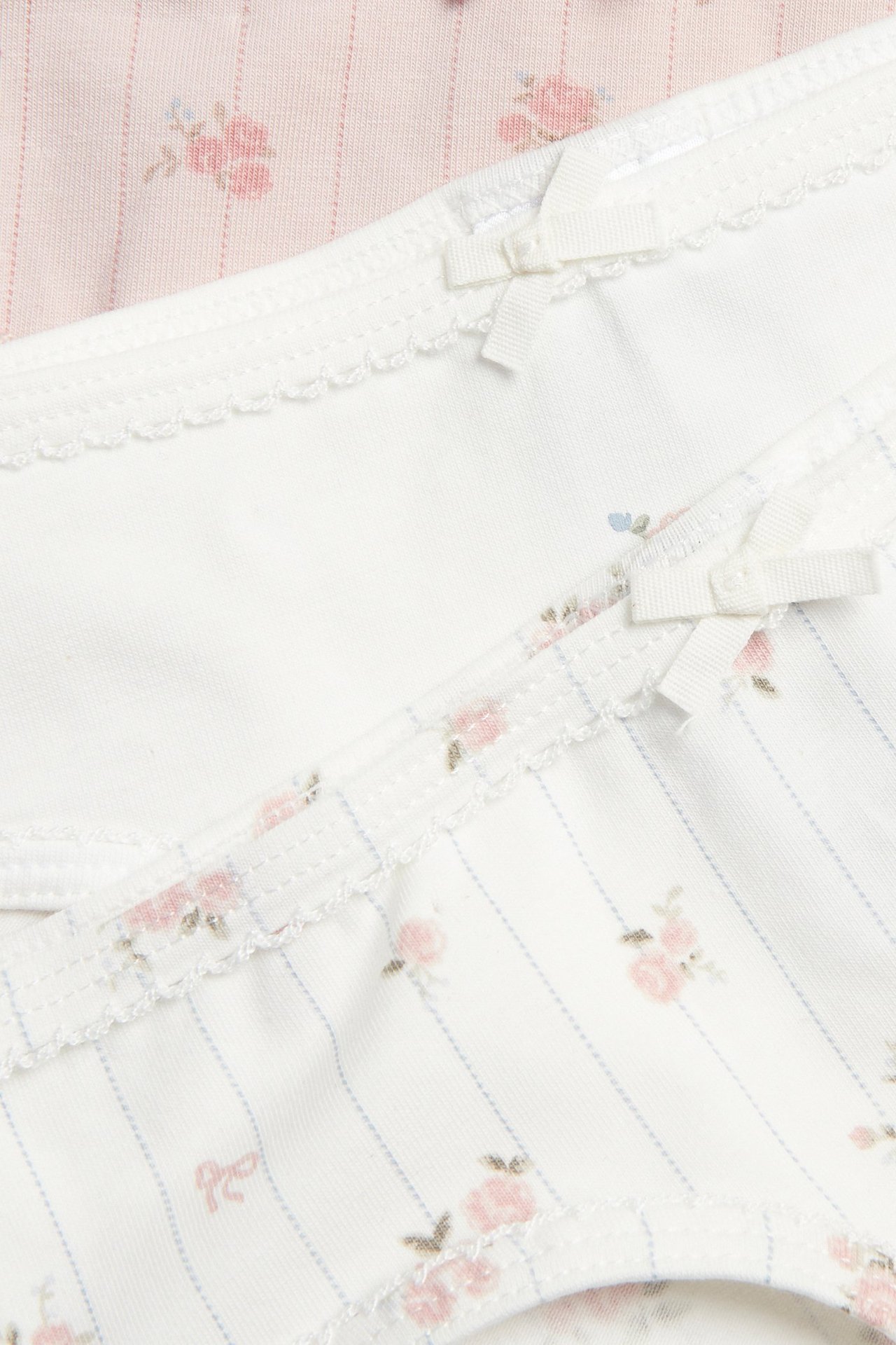 Zbliżenie: trzy bawełniane majtki w kwiaty, ecru w paski, ecru, różowe, z kokardkami i falbankami.