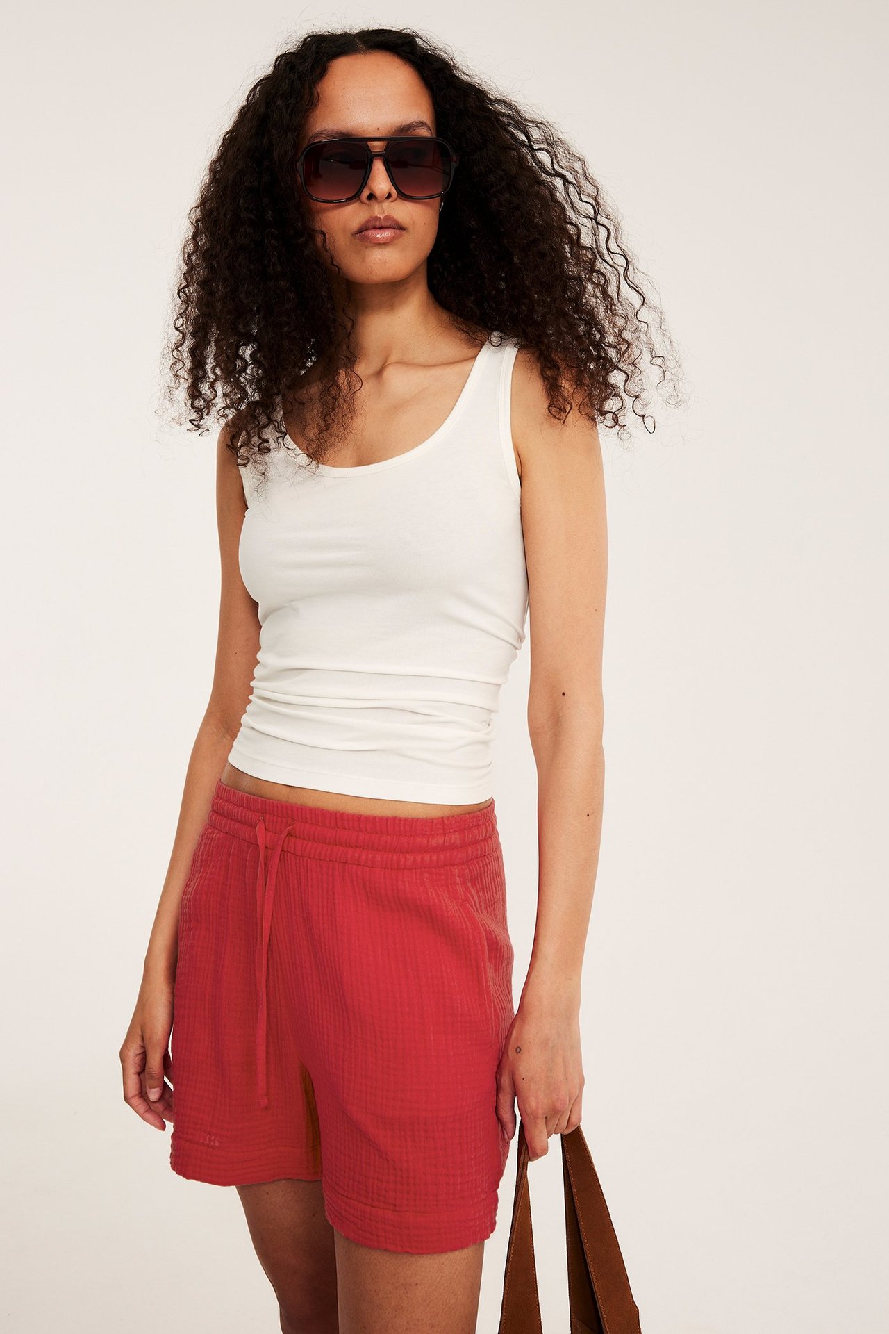 Framifrån en kvinna i röda strukturerade bomullsshorts, vit linnen, mörka solglasögon, håller en brun väska.