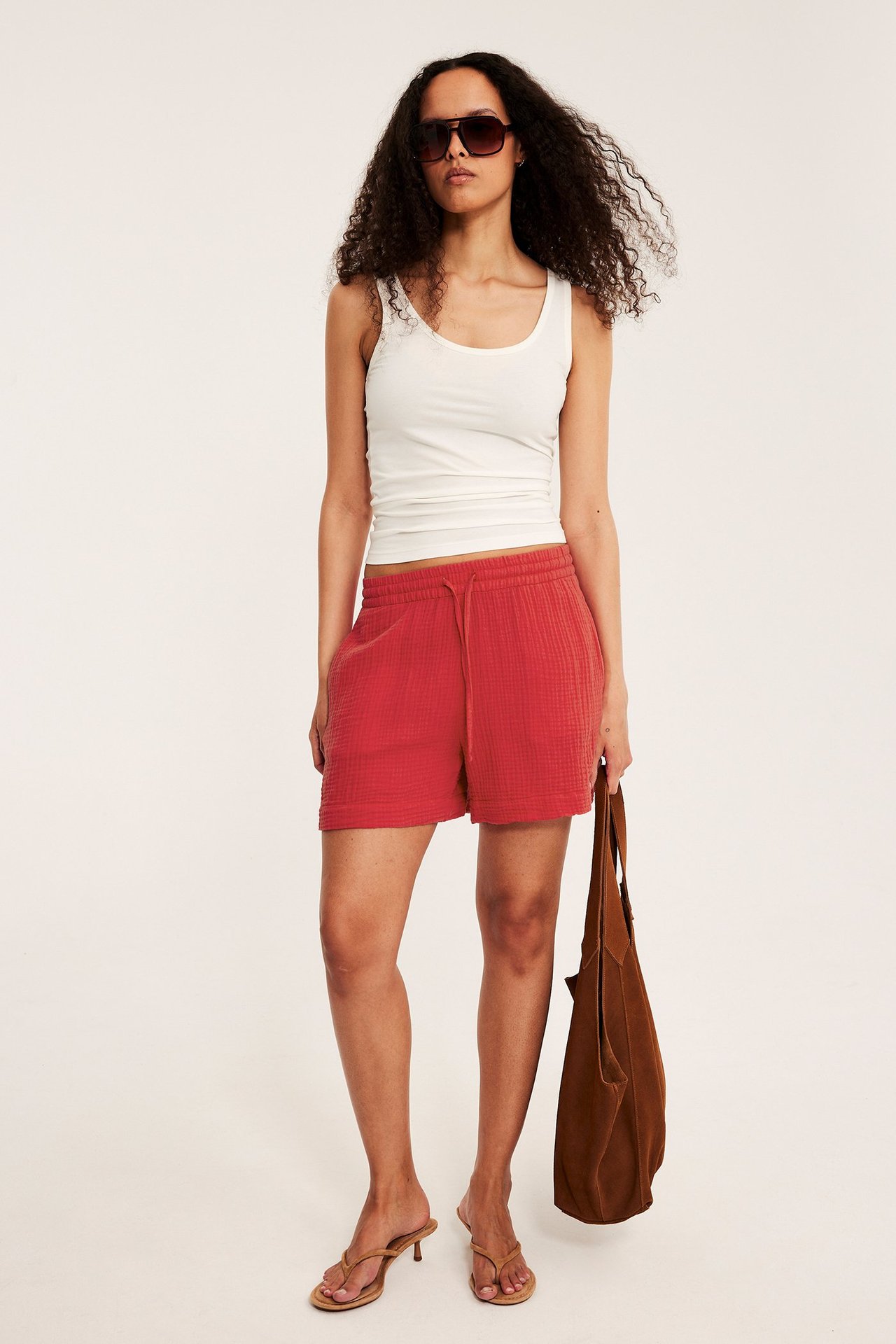 Edestä: nainen punaisissa shortsseissa, valkoisessa topissa, aurinkolaseissa, sandaaleissa, ruskea laukku kädessä.