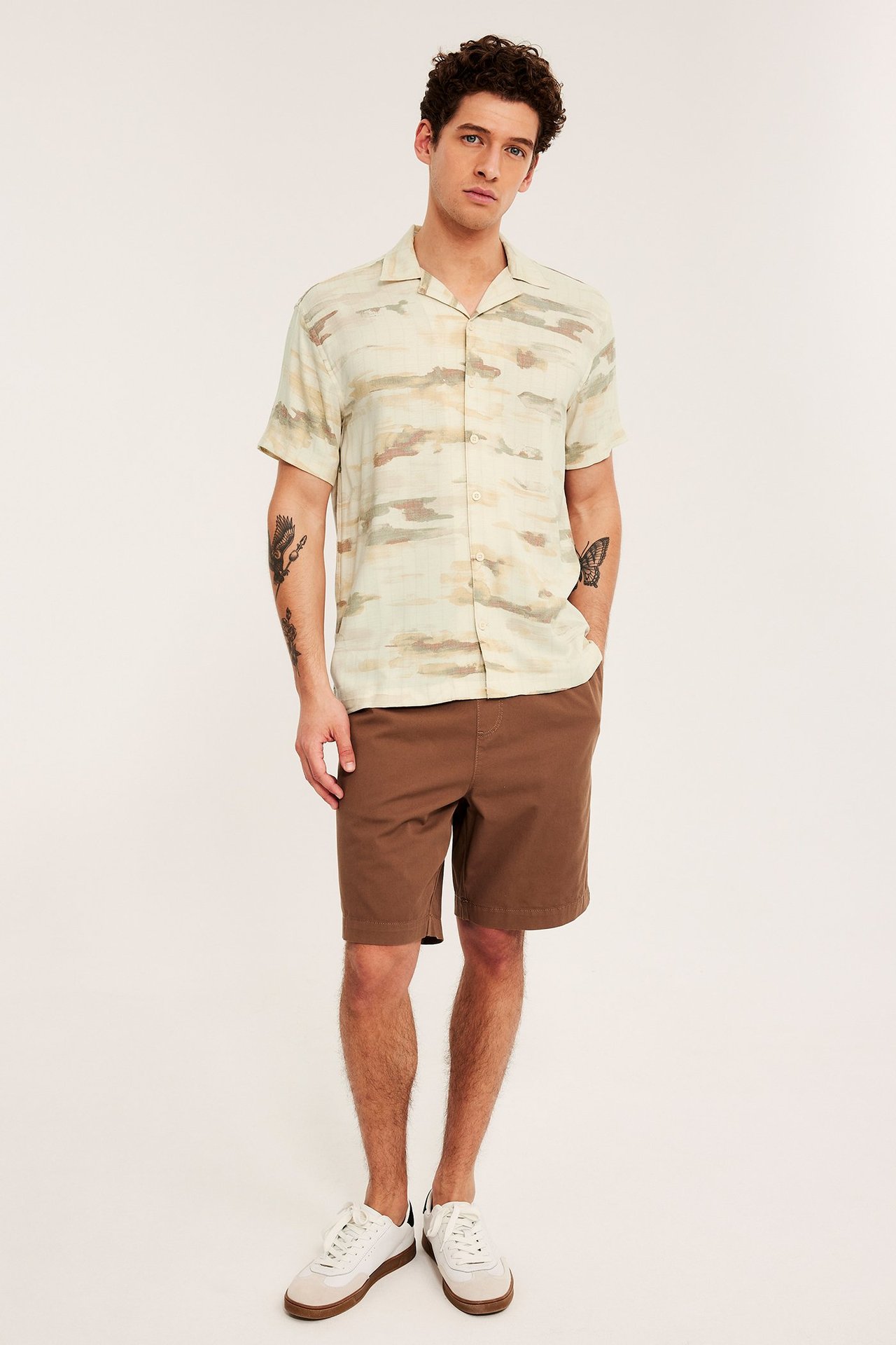 Framifrån: en man i beige mönstrad kortärmad resortsjorta, bruna shorts och vita sneakers.