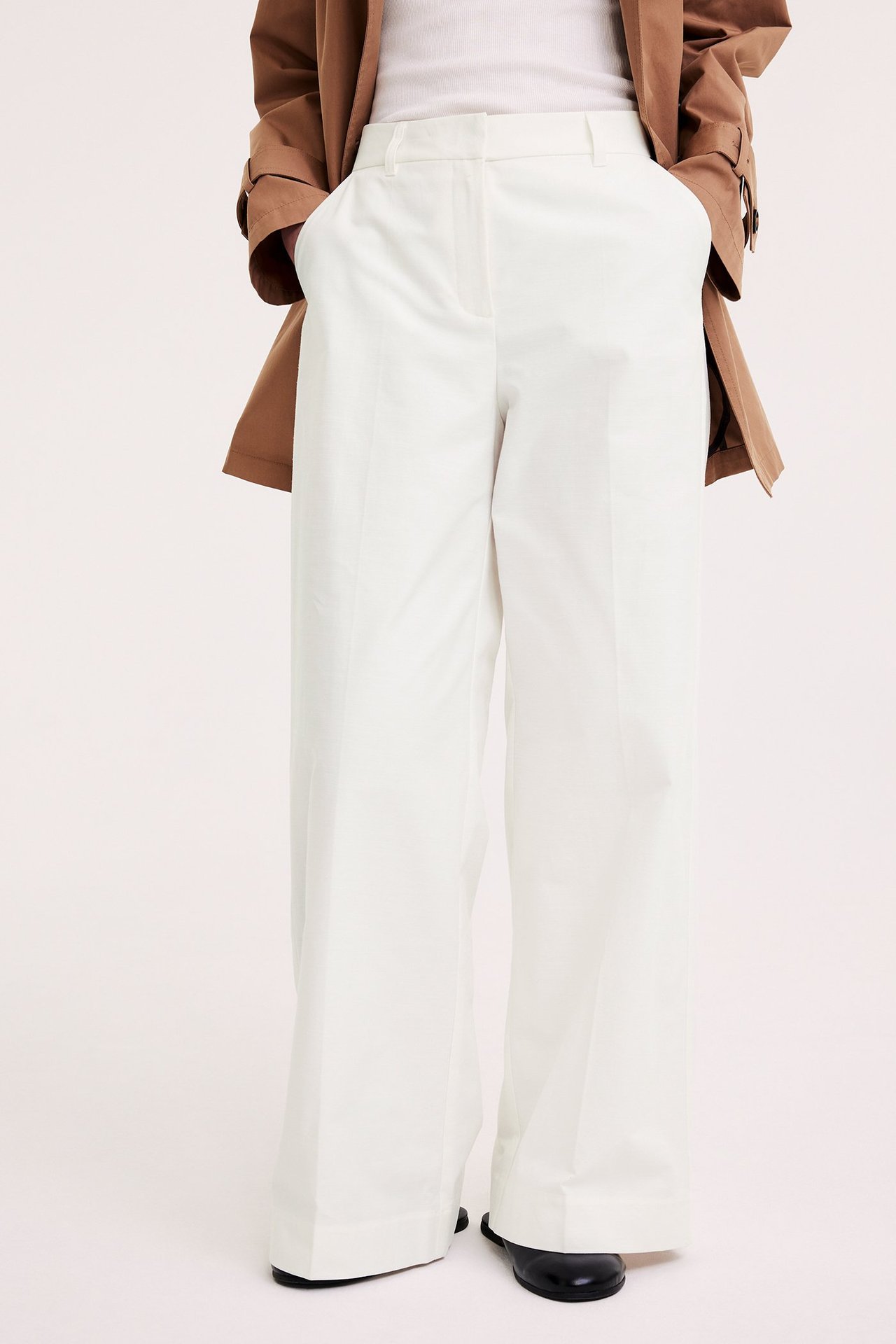 Framifrån: Kvinna i vita vida kostymbyxor med pressveck, vit topp, brun jacka och svarta skor.