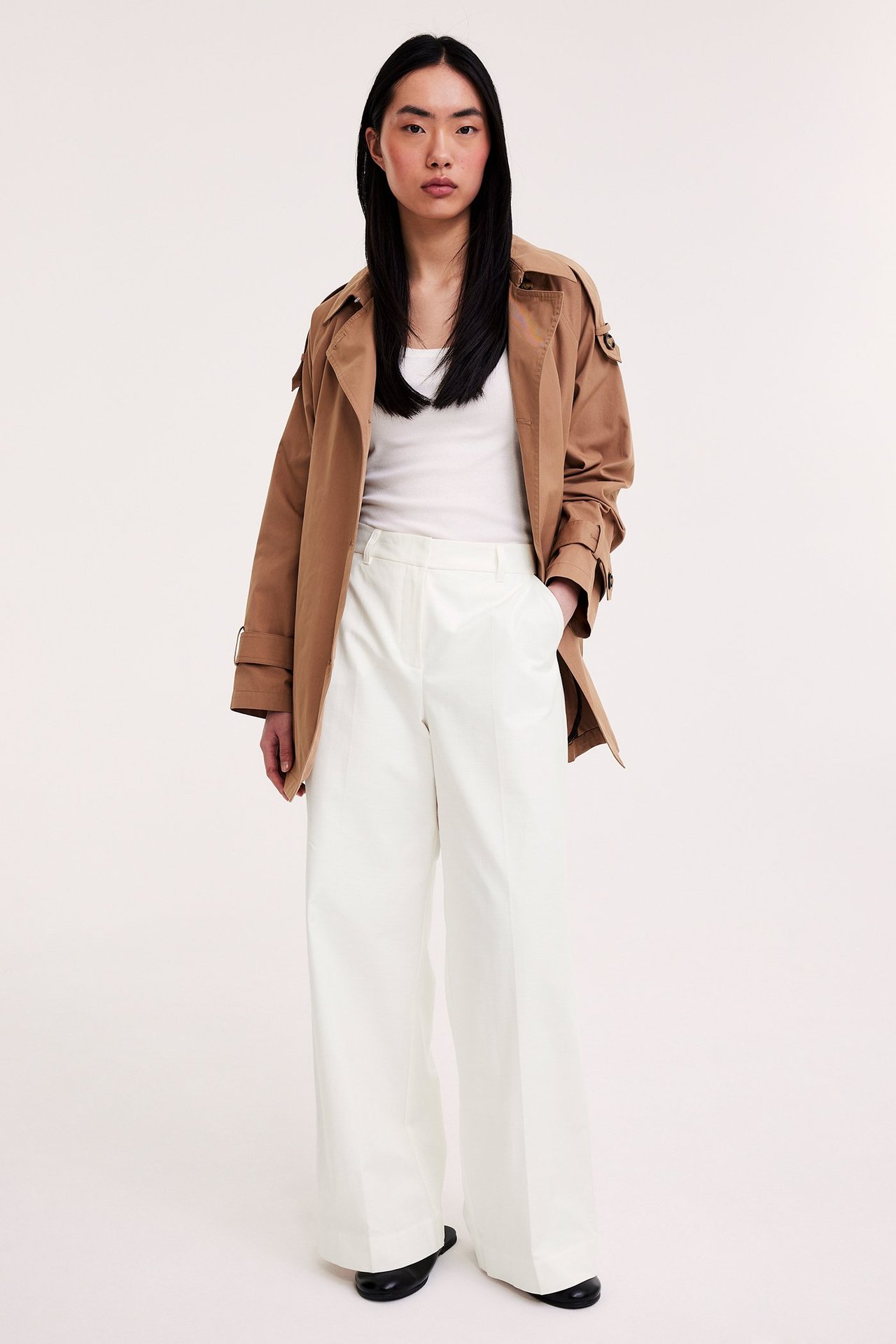 Edestäpäin: Nainen valkoisissa leveissä prässihousuissa, valkoisessa topissa ja vaaleanruskeassa trenssitakissa.