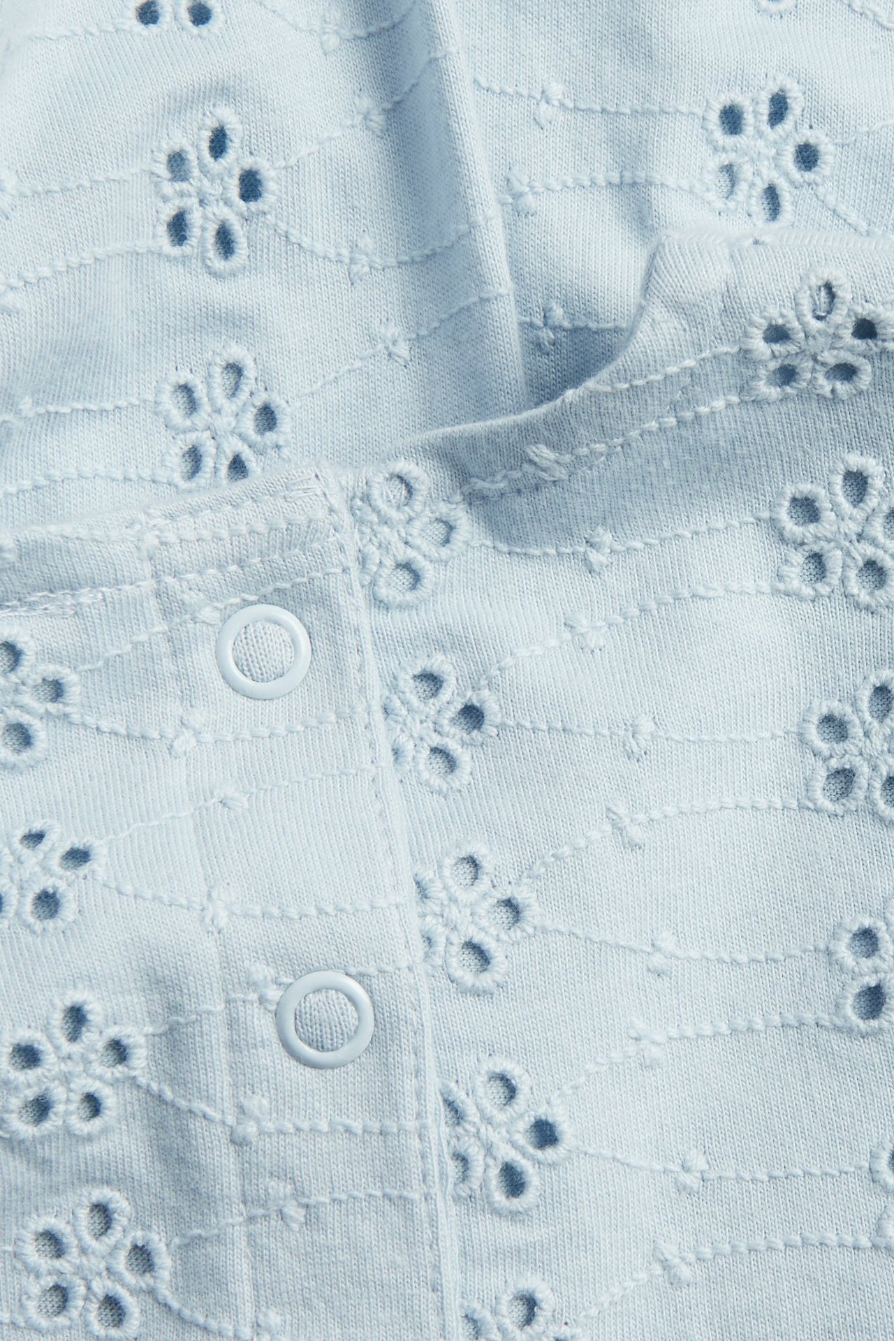 Nærbilde av en lyseblå babykjole i jersey med broderie anglaise blomstermønster og hvite trykknapper.