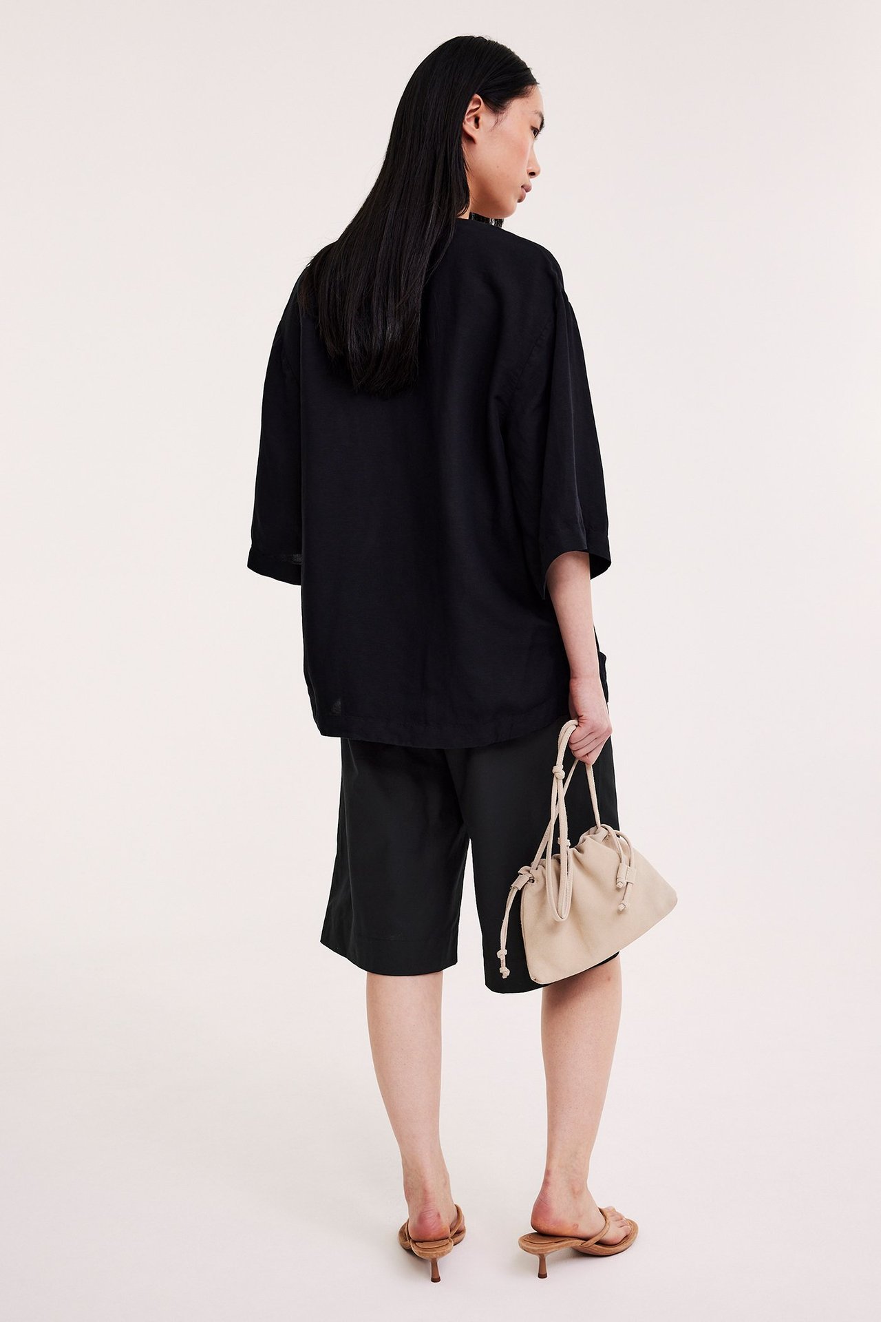 Ryggvy: Kvinna i svarta bomulls-bermudashorts, svart topp, bruna klacksandaler, håller en beige dragskoväska.