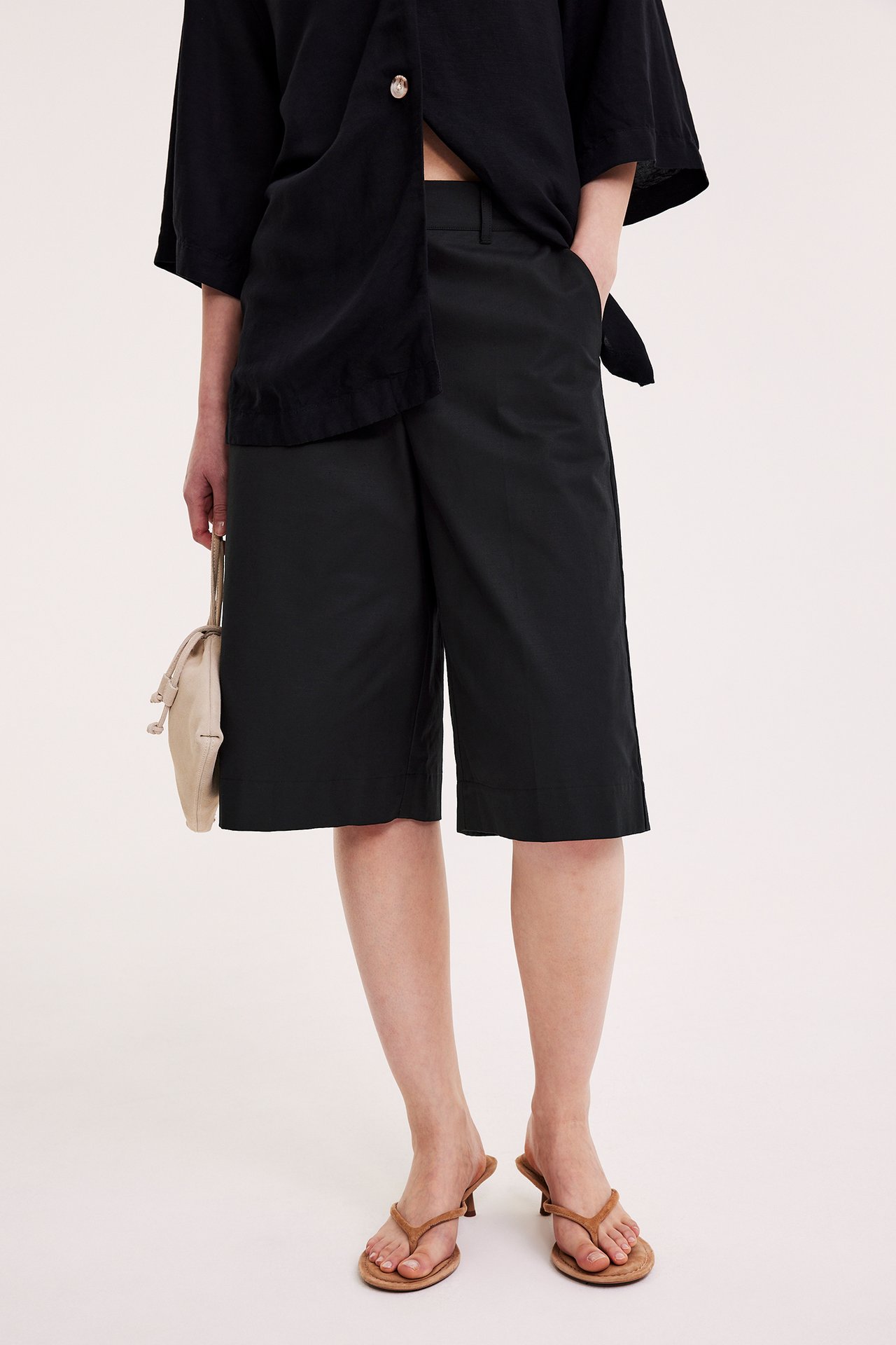 Framifrån, kvinna i svarta bomullsblandade bermudashorts, svart skjorta, bruna klacktofflor, och med en beige väska.