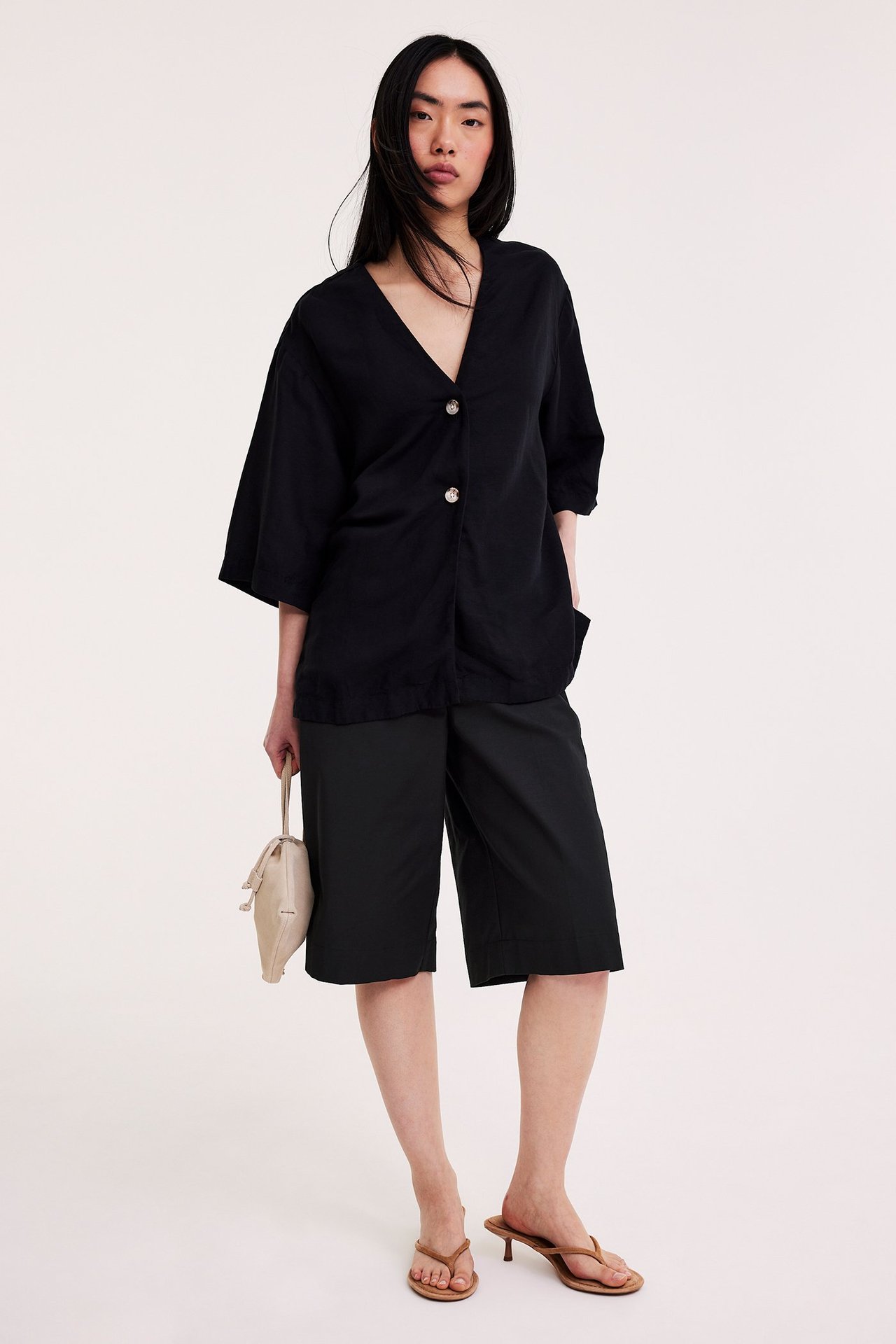 Framifrån, kvinna i svarta bermudashorts, svart V-ringad topp, bruna sandaler, håller ljusbeige handväska.