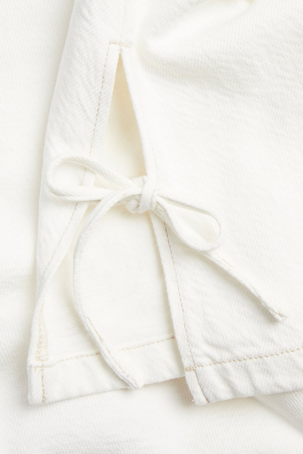 Nærbilde av en offwhite twilltopp med dekorative knytestropper i sidesømmen.
