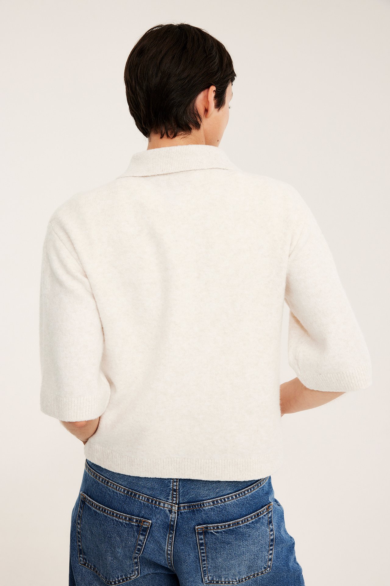 Ryggvy av en kvinna i en ljusbeige melerad stickad kofta med krage, kort ärm och blå jeans.