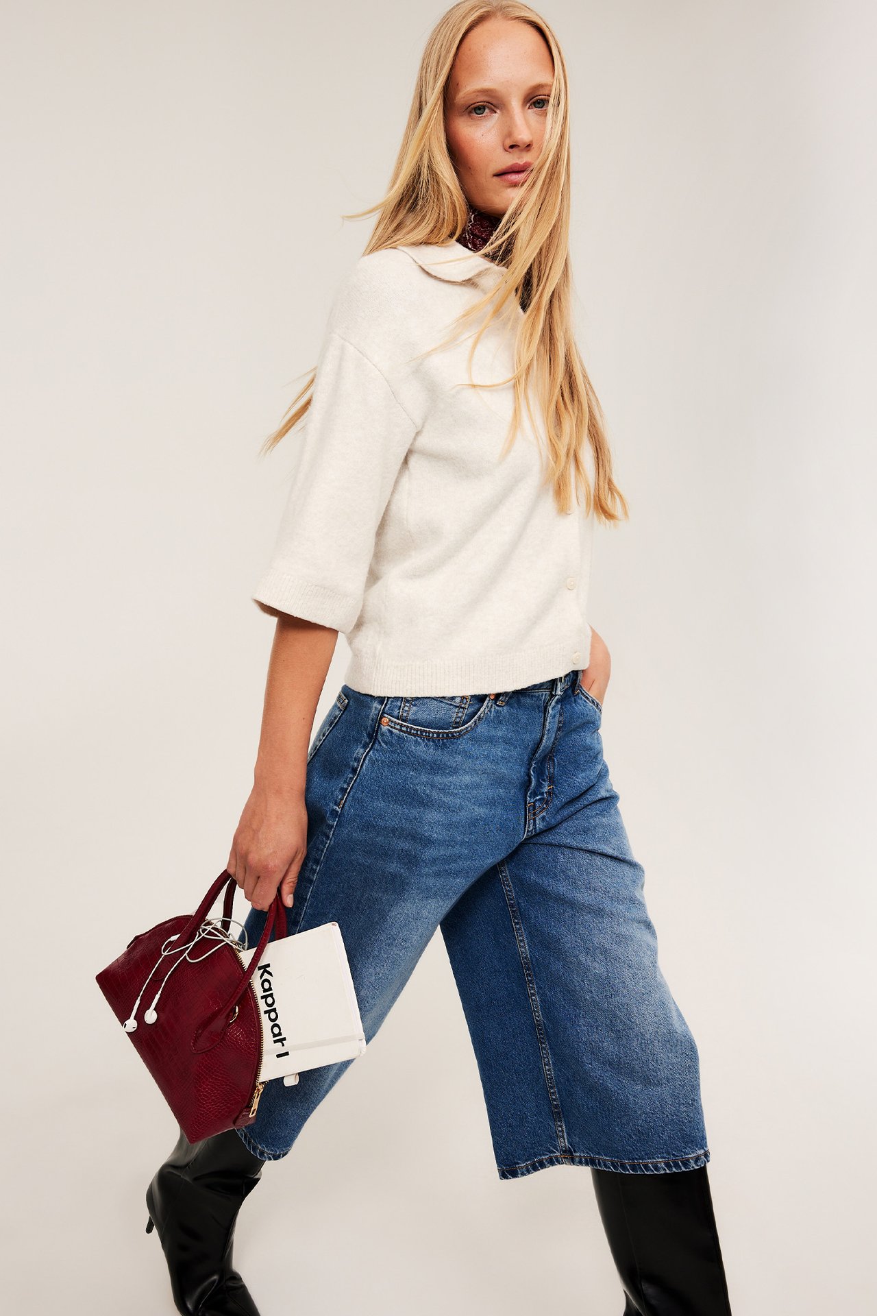 Framifrån: Ljusbeige kragad kofta, blå vida jeans, håller en mörkröd väska och vit liten påse.