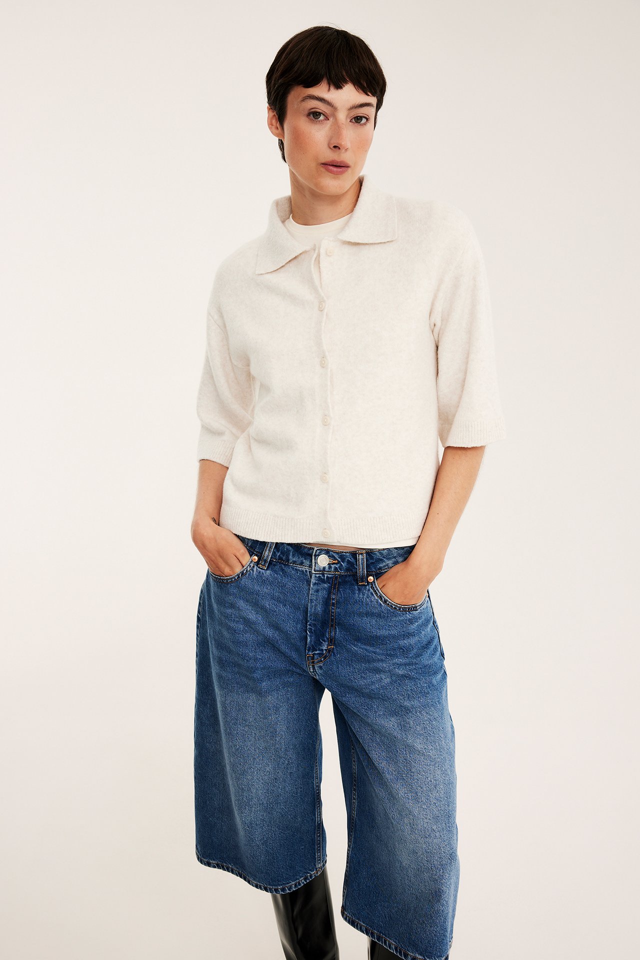 Framifrån: kvinna i ljusbeige melerad stickad kragekofta, vit topp och blå vida jeans.