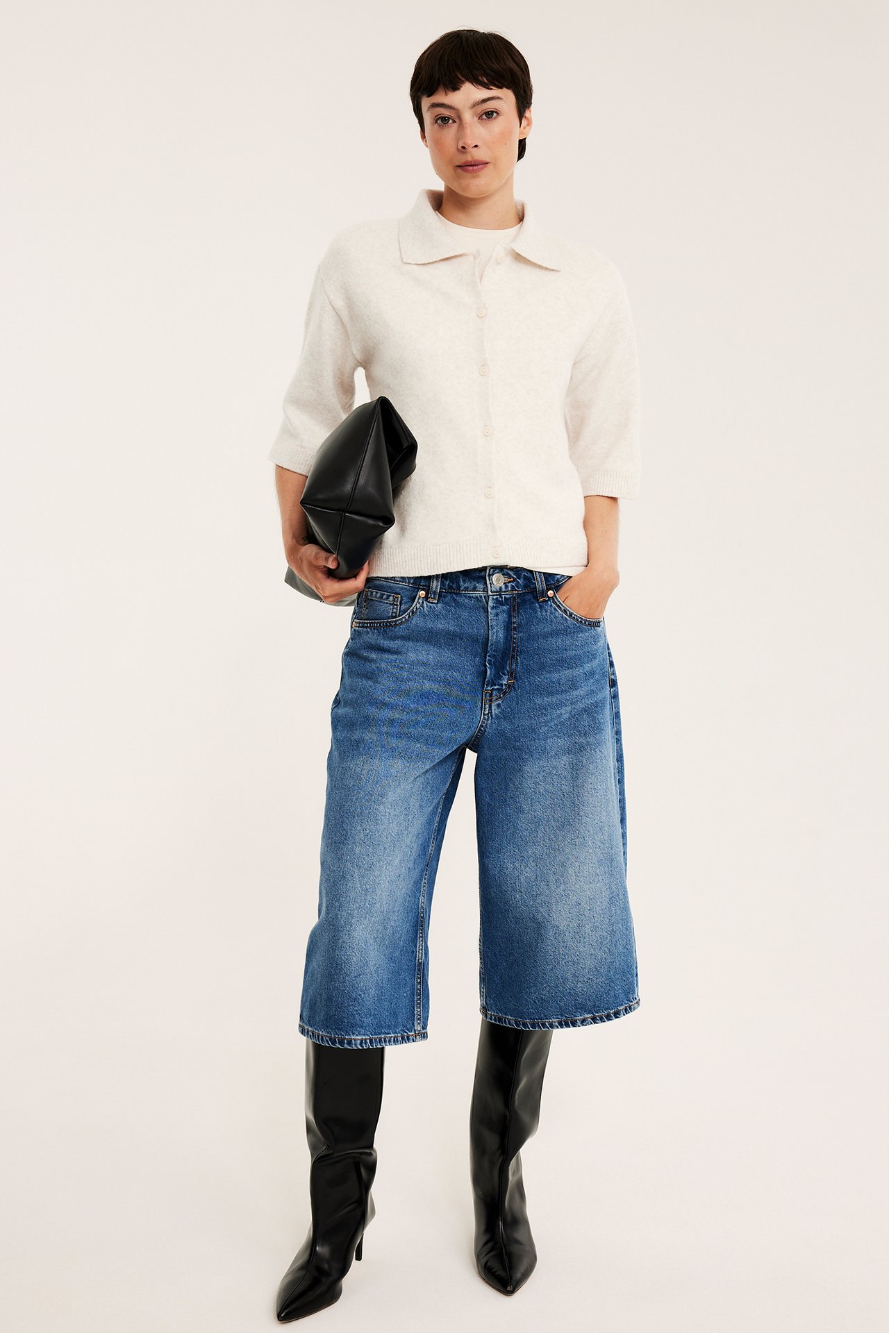 Framifrån: Kvinna i melerad ljusbeige stickad kofta, blå denim culottes, svarta stövlar, med svart clutchväska.