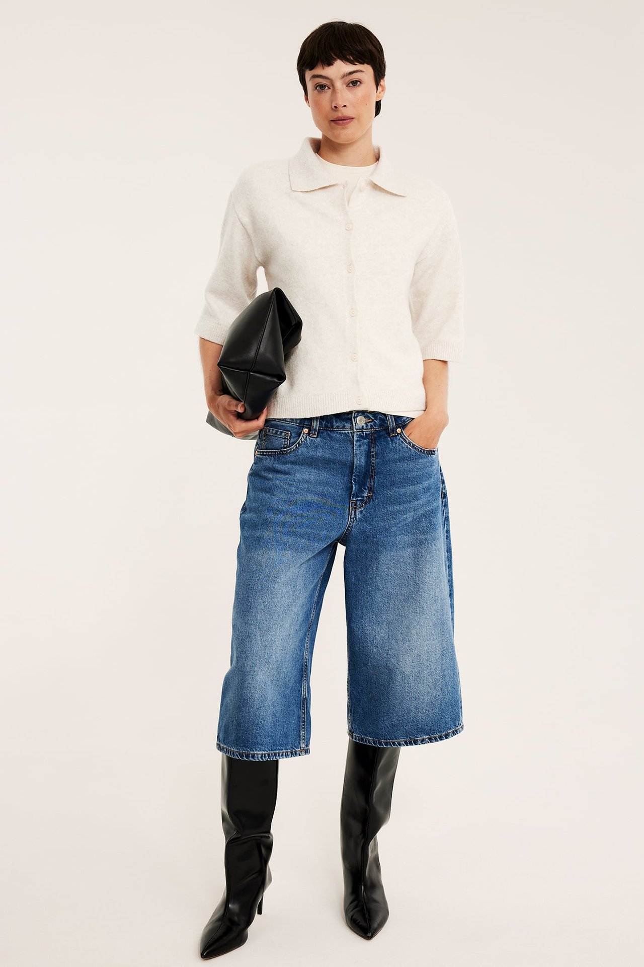 Framifrån: Kvinna i melerad ljusbeige stickad kofta, blå denim culottes, svarta stövlar, med svart clutchväska.