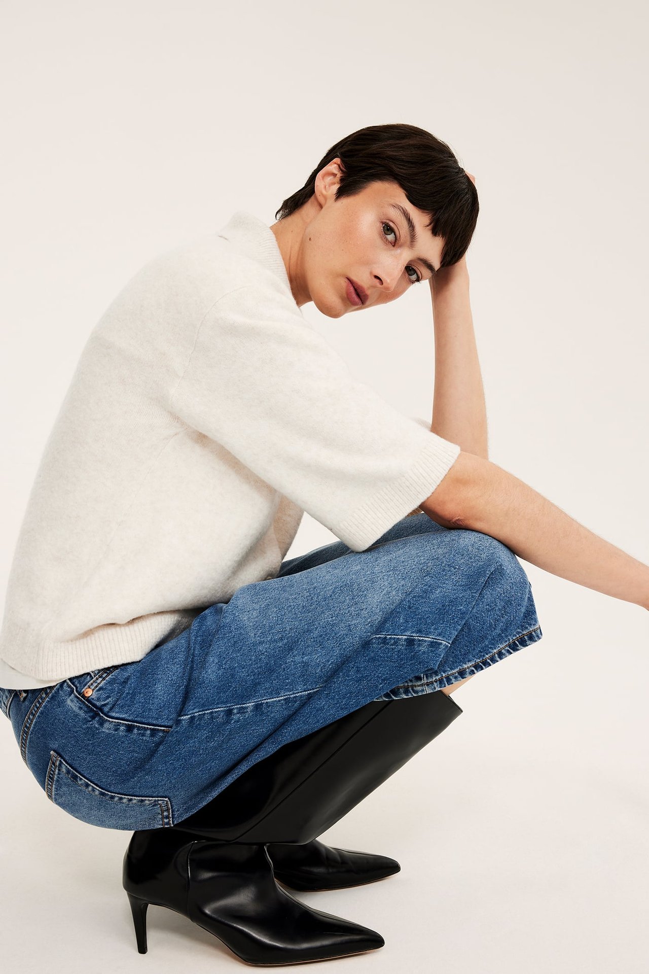 Från sidan, kvinna i ljusbeige melerad stickad kortärmad kofta med krage, blå jeans och svarta stövlar.