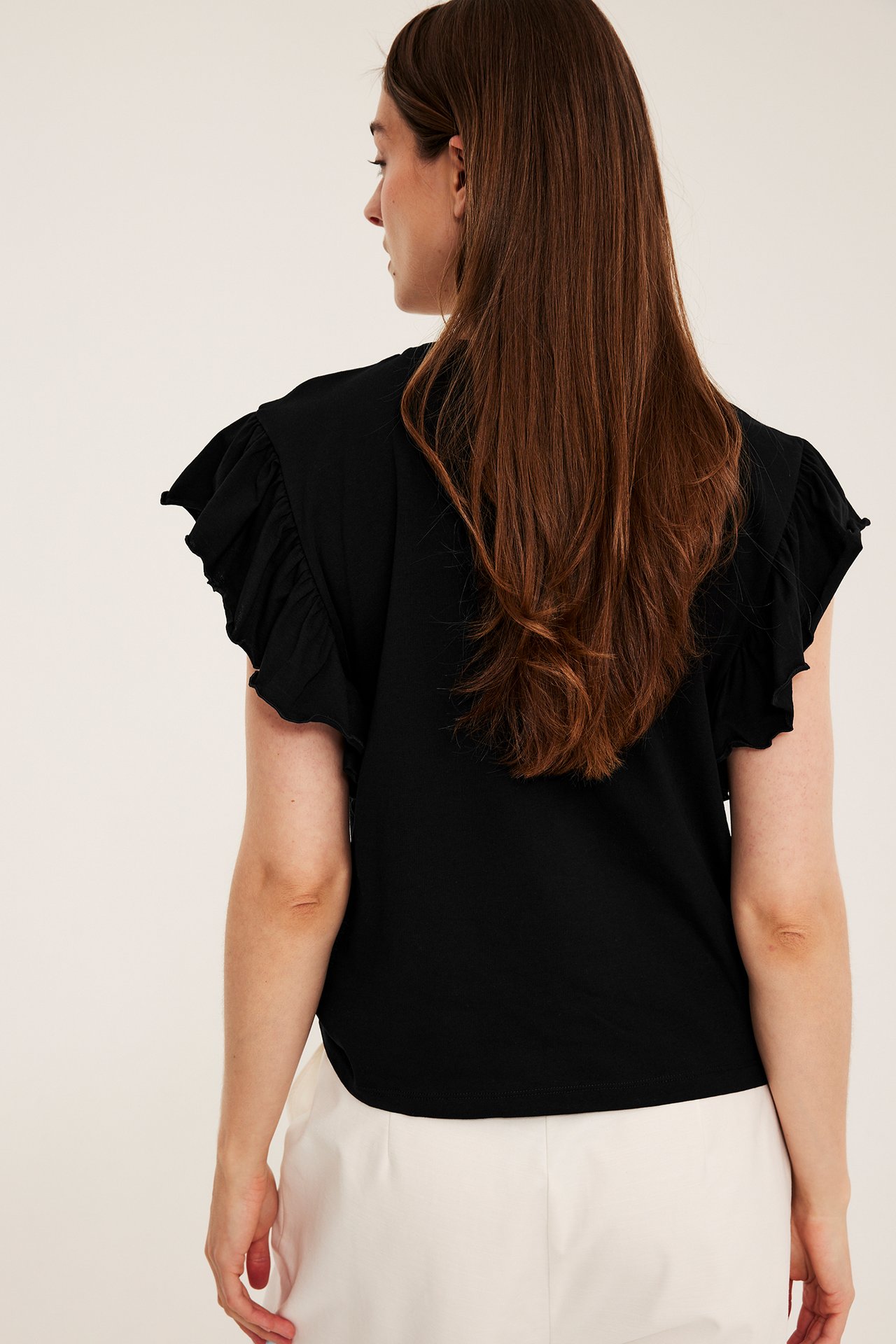 Bakfra sidevisning av kvinne i svart bomullsjerseytopp med rysjeermer og kremfargede bukser.