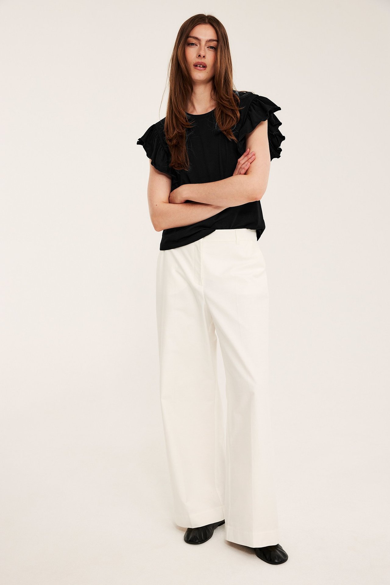Frontvy av en kvinna i svart bomullsjerseytopp med volangärm, vita vida byxor och svarta skor.