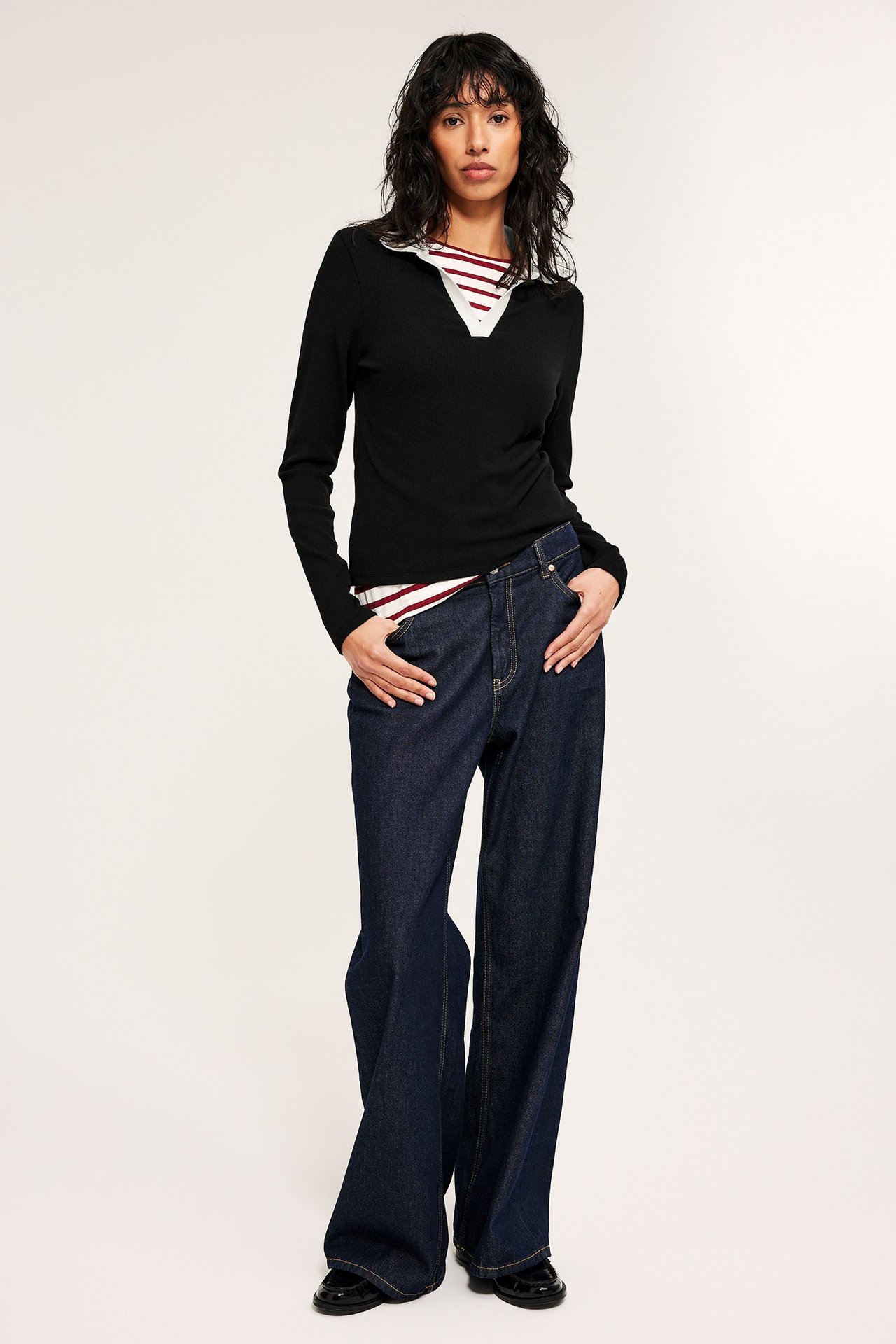 Edestä: Nainen mustassa istuvassa pitkähihaisessa kauluspaidassa, raidallinen paita, tummansiniset farkut, mustat loaferit.