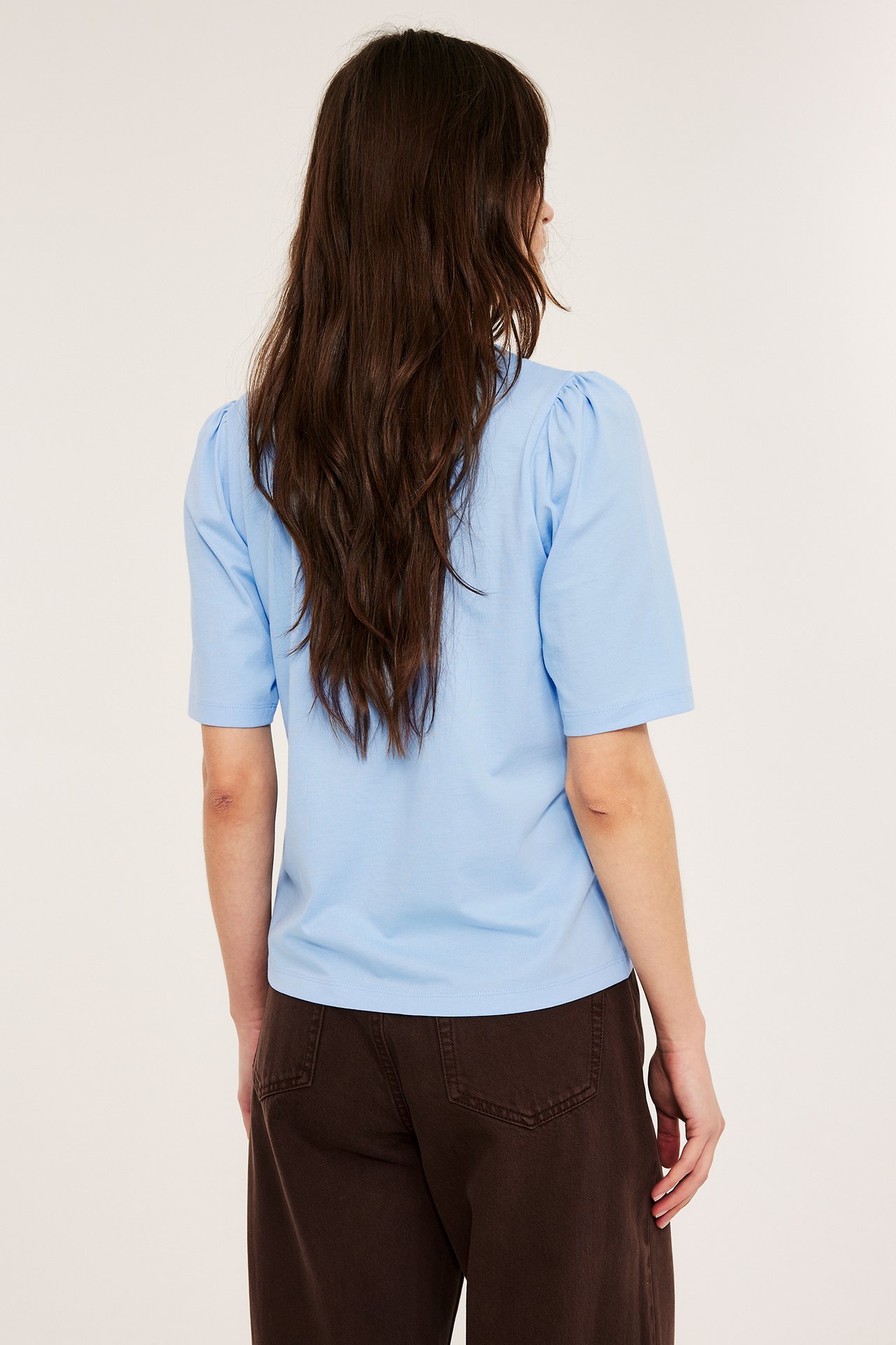 Ryggvisning av kvinne i lys blå bomullstopp med puffermer og mørkebrune bukser.