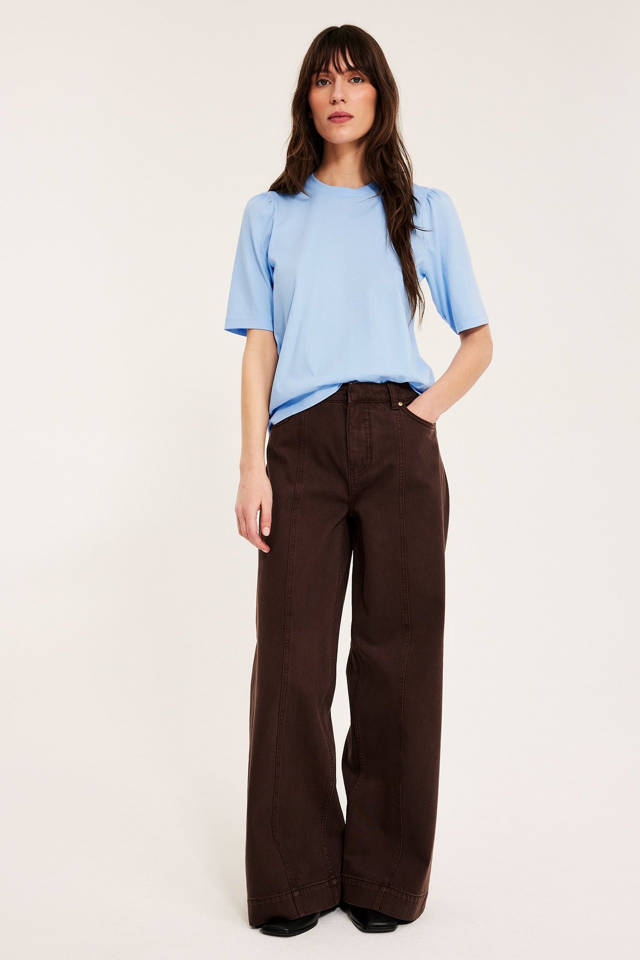 Forfra, kvinne i lyseblå bomullstopp med puffermer og mørkebrun vid bukse.