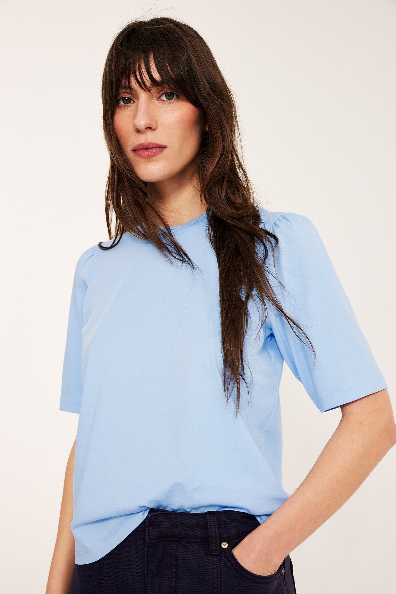 Frontbilde av kvinne i lyseblå bomullstopp med puffermer og mørkeblå bukser.