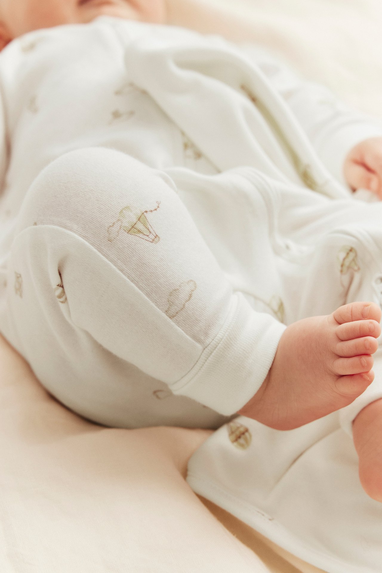 Lähikuva: vauva yllään luonnonvalkoinen puuvillaromper, jossa ilmapalloja, vetoketju ja käännettävät resorit.