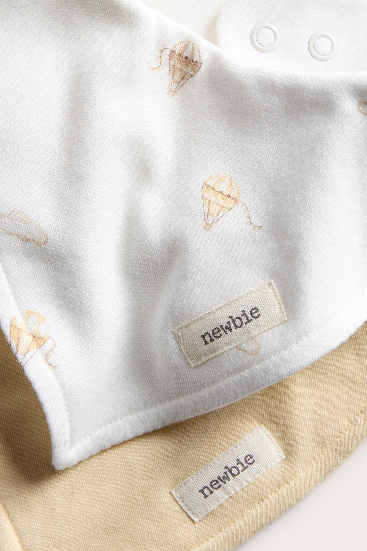 Närbild av två babybomulls-dregglisar: en offwhite med luftballonger och tryckknappar, en enfärgad ljusbeige.