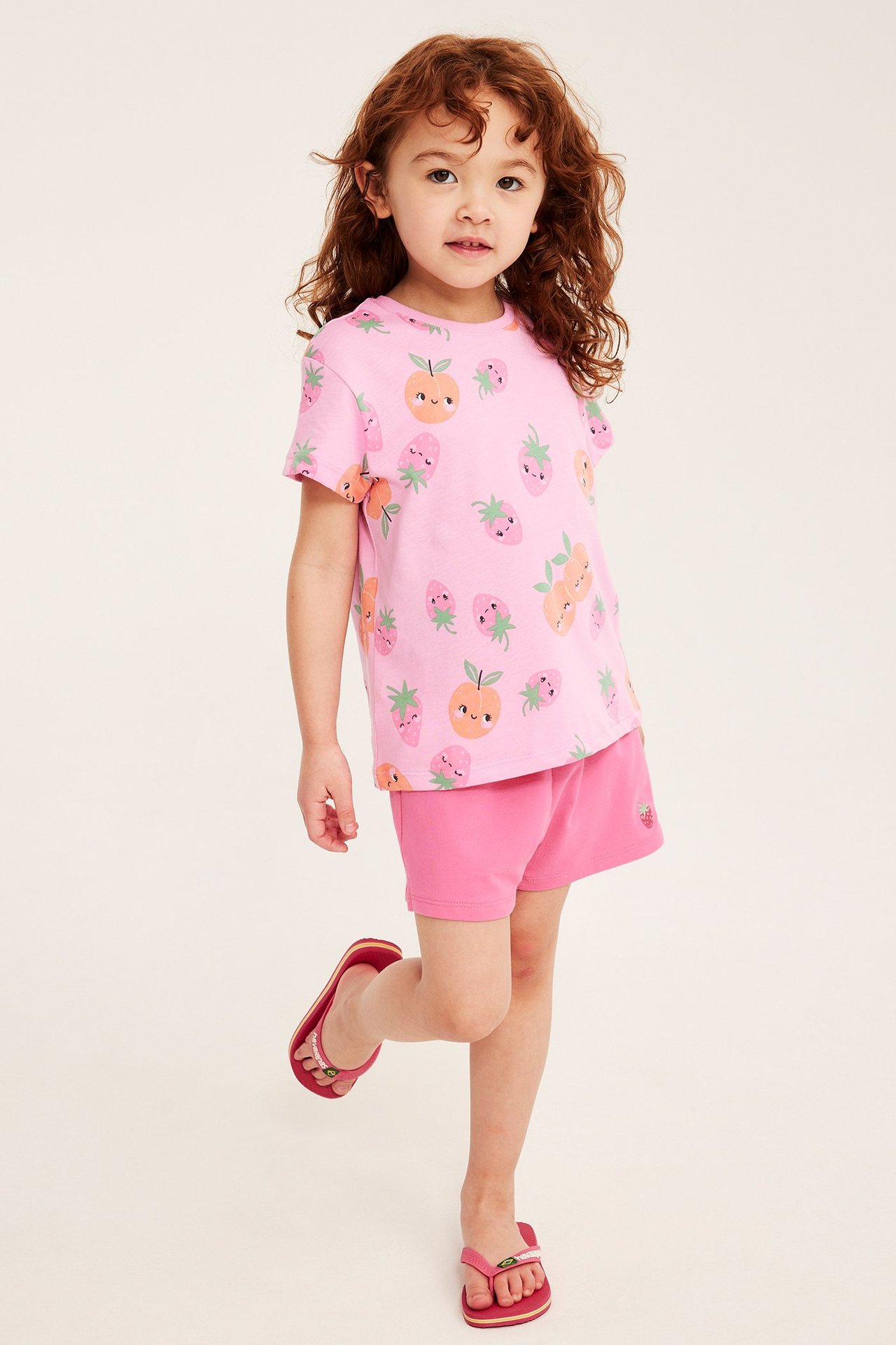Frontbilde av et barn i rosa shorts med jordbærbroderi, en lyserosa t-skjorte med fruktmønster og røde flip-flops.