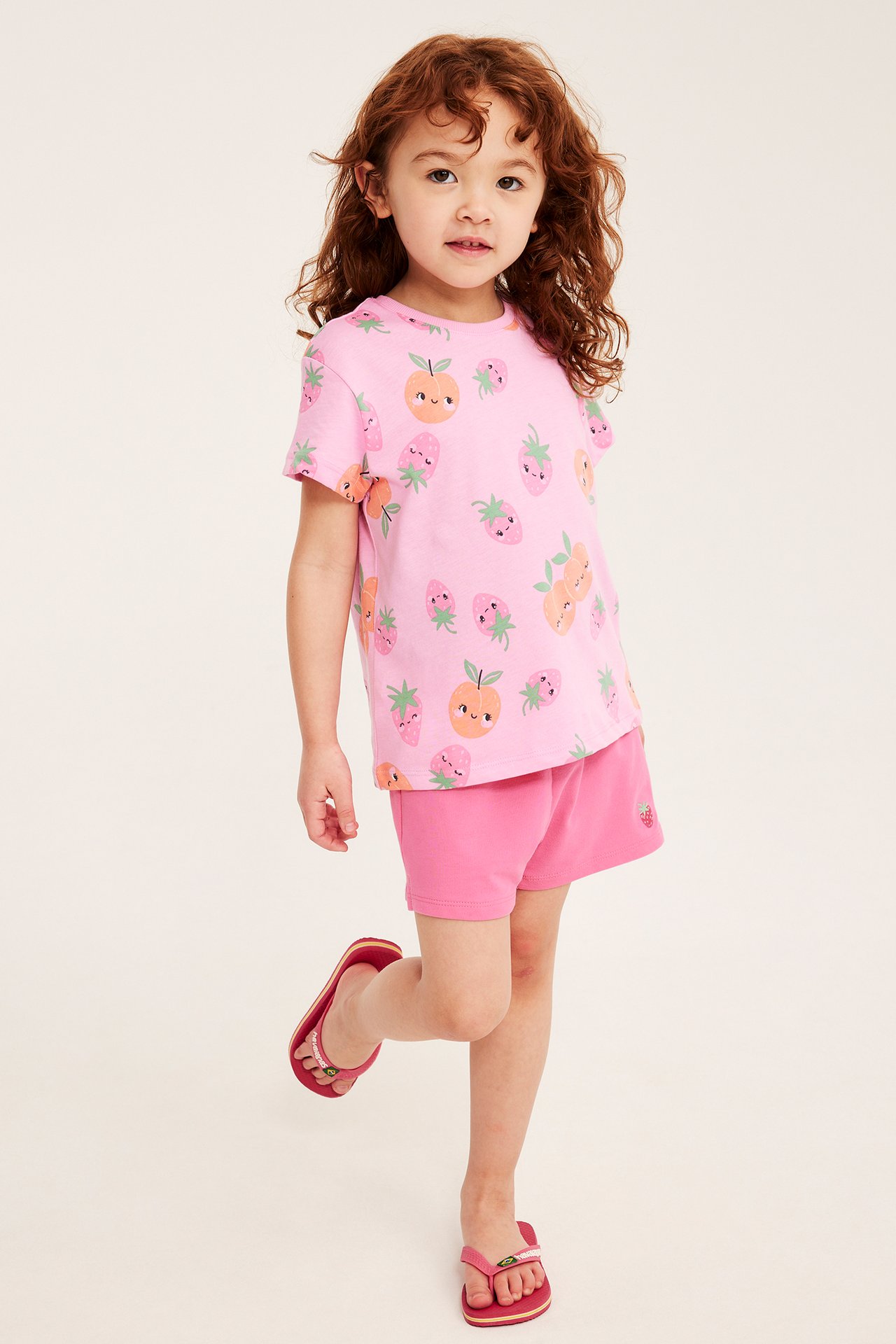 Widok z przodu dziecka w różowych szortach z haftem truskawkowym, jasnoróżowej koszulce w owoce i czerwonych klapkach.