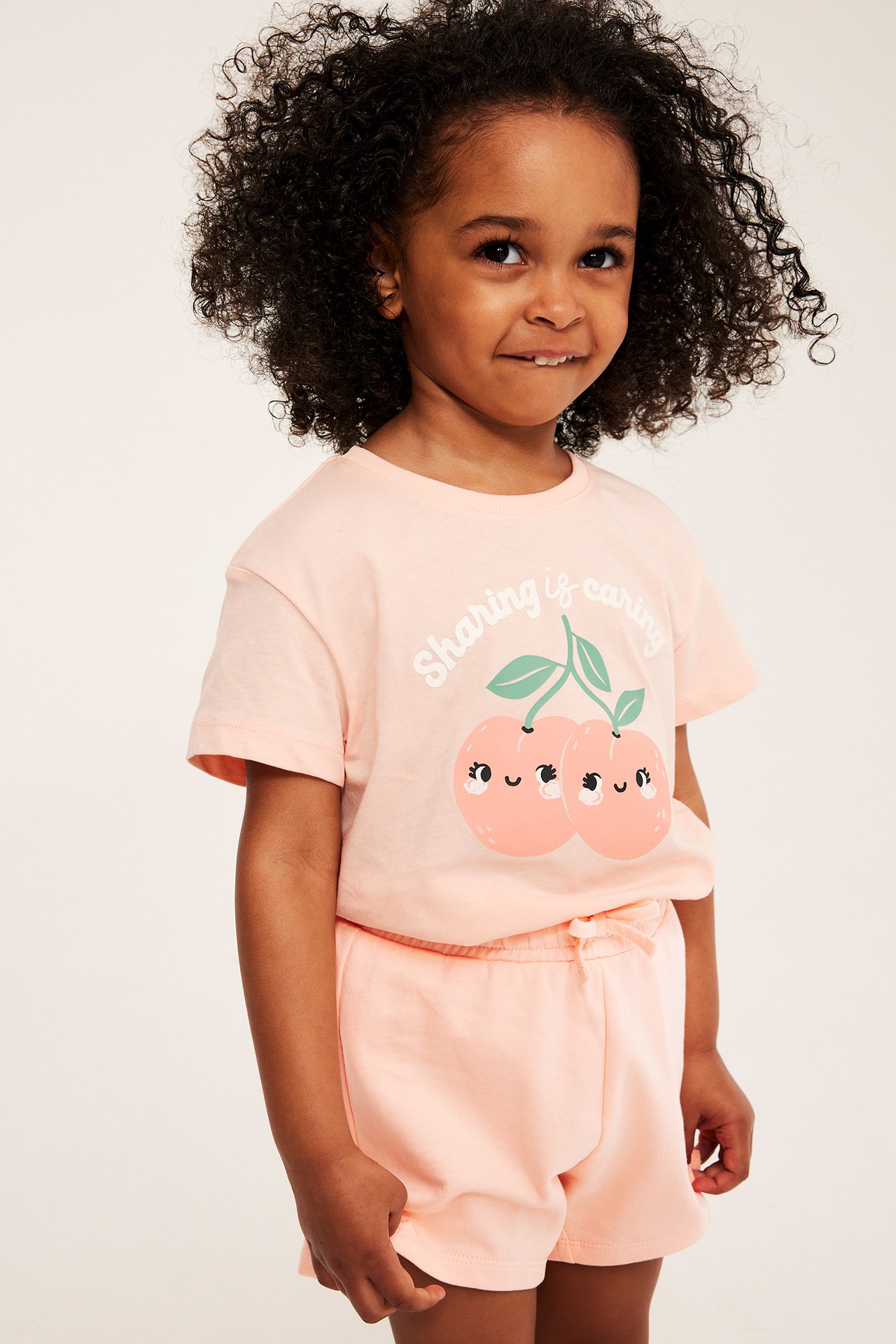 Edestäpäin, lapsi yllään korallinväriset shortsit rusetilla ja yhteensopiva t-paita kirsikkakuviolla.